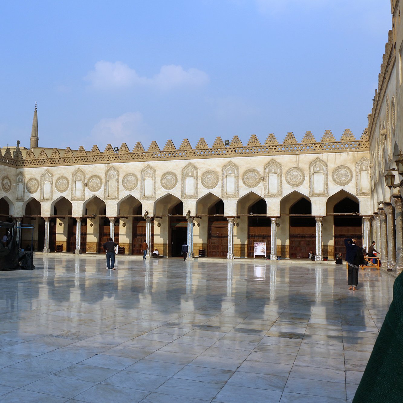 الصورة تظهر ساحة كبيرة داخل معلم تاريخي، يحتمل أن يكون مسجدًا أو جامعة إسلامية. الساحة محاطة بأقواس زخرفية وبهياكل عالية تظهر الهندسة المعمارية الإسلامية التقليدية. الأرض مغطاة ببلاط لامع يعكس ضوء السماء، مما يضفي جمالية خاصة على المكان. في الخلفية، يمكن رؤية قبة وجدران مزخرفة، بالإضافة إلى بعض الأشخاص الذين يتجولون في المكان. السماء زرقاء مع بعض الغيوم، مما يضيف لمسة من الهدوء إلى المشهد.