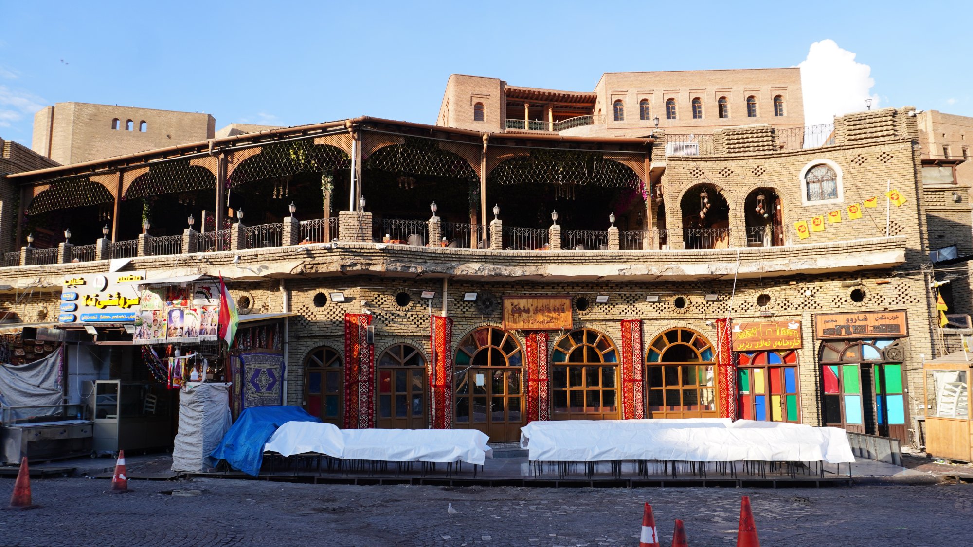 The image depicts a beautifully constructed building featuring intricate architectural details, likely in a historic or culturally significant area. The facade showcases ornate designs, colorful window frames, and a balcony adorned with wooden railings. The ground level appears to have some protective coverings, possibly due to ongoing renovations or maintenance. Surrounding the building, there are orange traffic cones indicating restricted access in that area. The overall ambiance suggests a blend of tradition and vibrant community life.