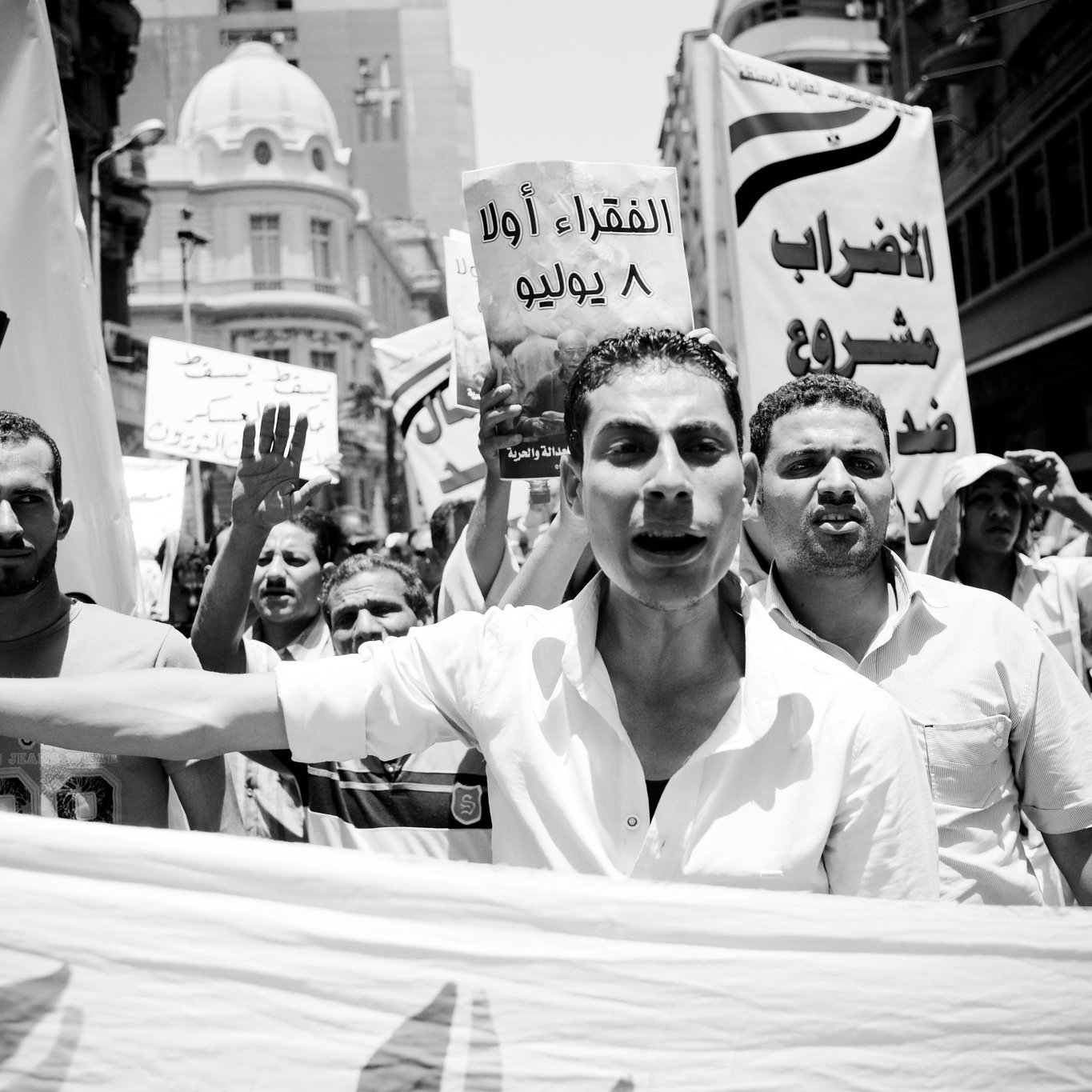 L'image montre une manifestation animée en noir et blanc. Des participants tiennent des pancartes et expriment leur mécontentement. Au premier plan, un homme semble parler avec passion, tandis que d'autres personnes en arrière-plan lèvent les bras et crient. Les banderoles sont en arabe, indiquant des revendications sociales ou politiques. L'ambiance est dynamique et engagée.