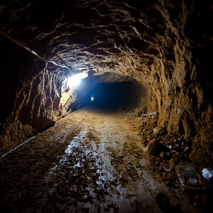 Cette image montre un tunnel souterrain. Les parois sont irrégulières et terreuses, indiquant qu'il a été creusé à la main ou à l'aide d'outils simples. Le sol est recouvert de terre, et une source de lumière artificielle éclaire l'intérieur, créant un contraste entre l'obscurité du tunnel et la lumière. À droite, on peut apercevoir des déchets éparpillés, ce qui suggère que l'endroit a été utilisé pour un certain temps. Le tunnel semble relativement étroit et évoque une atmosphère de mystère ou d'aventure.