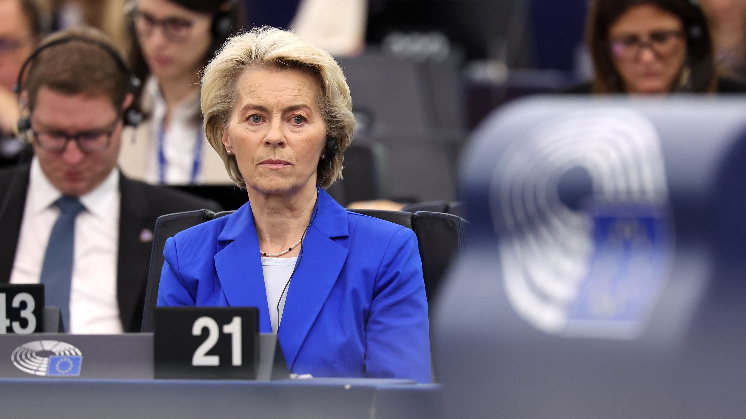 The image features a formal setting, likely a parliamentary or governmental session. A woman with short, light-colored hair and dressed in a blue blazer is seated prominently, looking serious and focused. She appears to be attending an important meeting or discussion, with microphones and attendees in the background, indicating a formal and possibly political environment. Others in the background are also engaged, wearing headsets and taking notes.