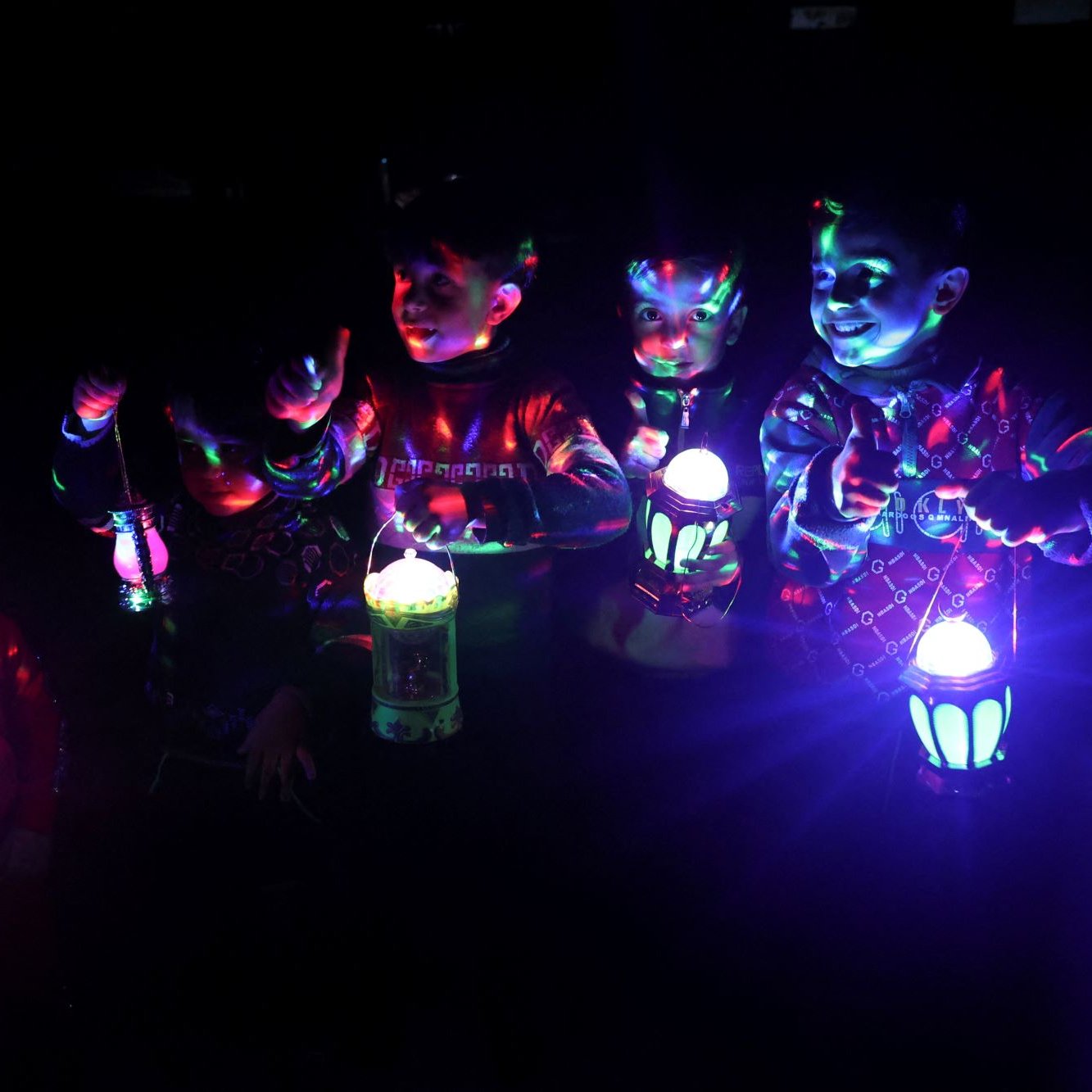 Nell'immagine si possono vedere sei bambini che si trovano in un ambiente buio, illuminati da luci colorate che proiettano effetti dinamici. Ognuno dei bambini tiene una lanterna che emette una luce vivace e colorata. I piccoli sembrano divertirsi, sorridendo e interagendo tra di loro. Le loro espressioni trasmettono gioia e entusiasmo, creando un'atmosfera festosa. Le luci colorate e le ombre contribuiscono a rendere l'immagine molto suggestiva.