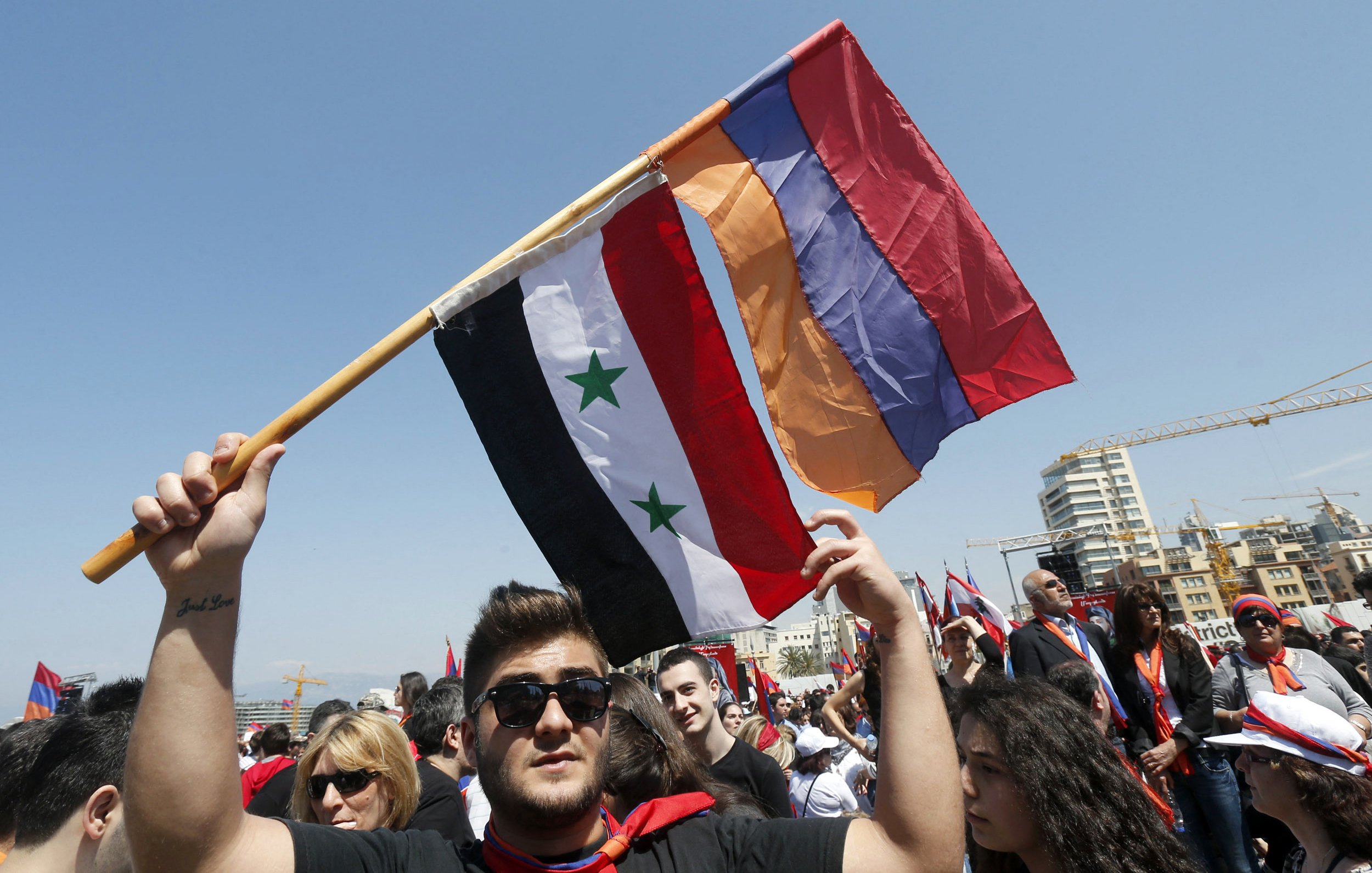 L'image montre un groupe de personnes participant à une manifestation ou un rassemblement. Un homme au premier plan tient fièrement trois drapeaux : le drapeau syrien, un drapeau avec trois étoiles vertes sur fond noir et rouge, ainsi qu'un drapeau arménien. L'ambiance semble festive et engagée, avec de nombreuses personnes en arrière-plan, probablement partageant des sentiments communs ou soutenant une cause. Le ciel est clair, ce qui ajoute à l'énergie de l'événement.