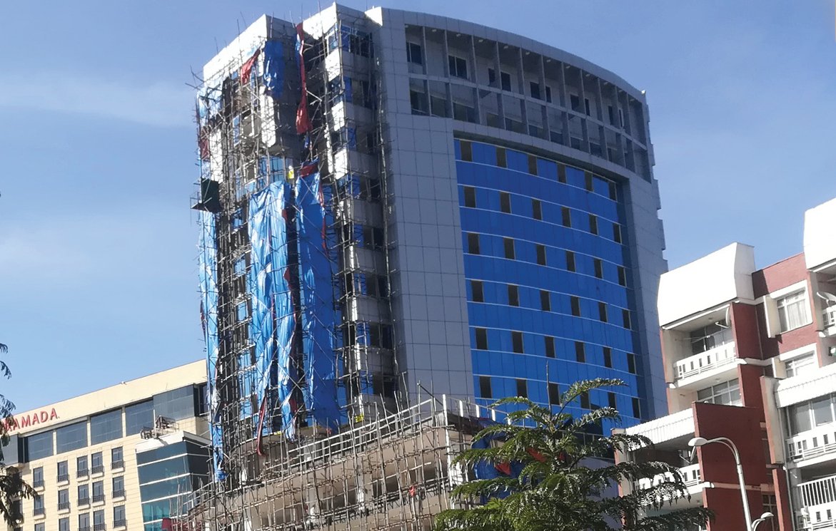 L'image montre un bâtiment en construction. Il s'agit d'une structure de plusieurs étages, avec une façade partiellement terminée, affichant des panneaux de verre de couleur bleue et d'autres en blanc. Le bâtiment est entouré d'échafaudages en métal, et des bâches bleues sont visibles sur certains côtés. À proximité, on aperçoit d'autres bâtiments, probablement de bureaux ou commerciaux, et un ciel bleu en toile de fond. La végétation est également présente au premier plan, ajoutant une touche de verdure à l'environnement urbain.