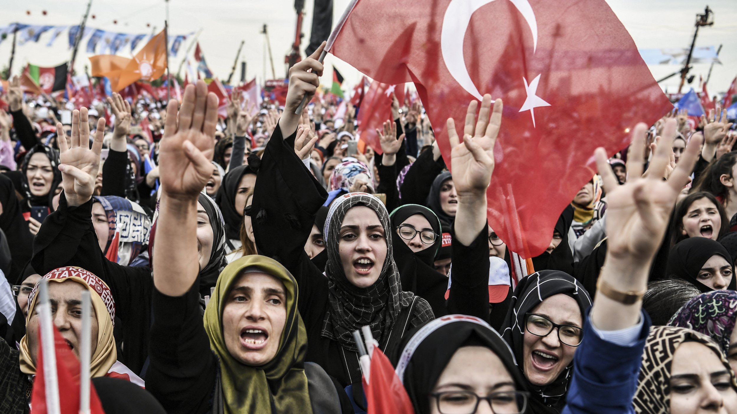 این تصویر نمایی از جمعیتی از زنان را نشان می‌دهد که در یک تجمع یا راهپیمایی شرکت کرده‌اند. آنها پرچم‌های ترکیه را در دست دارند و با شور و هیجان شعار می‌زنند. بیشتر زنان در عکس با حجاب دیده می‌شوند و برخی از آنها با دست علامت پیروزی نشان می‌دهند. فضای عمومی و اجتماعی این تصویر بسیار فعال و پر جنب و جوش به نظر می‌رسد.