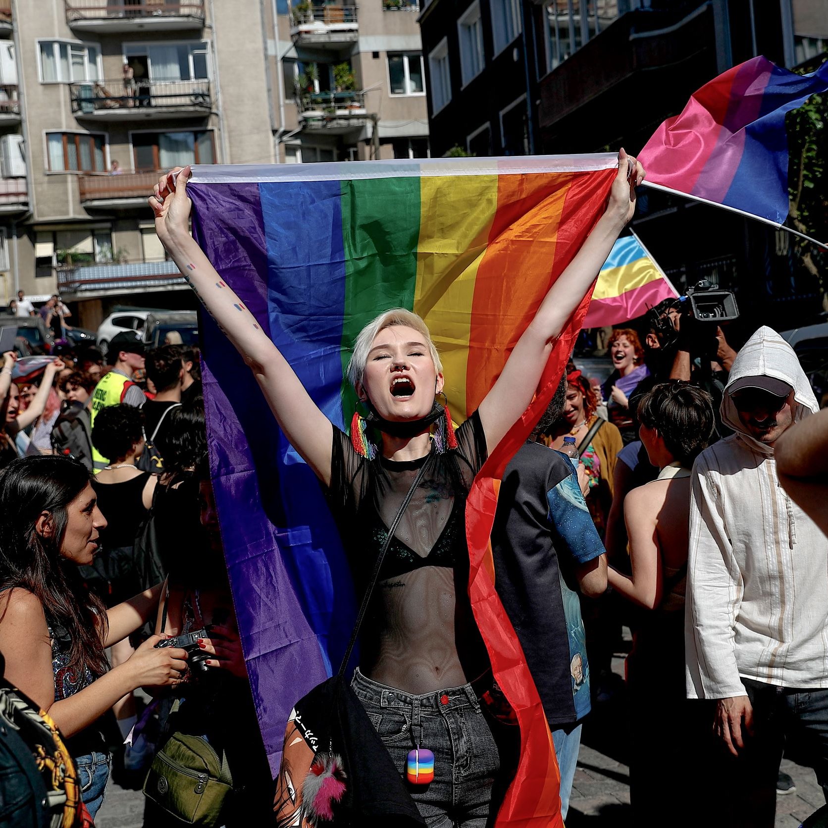 L'image montre une scène dynamique d'une marche ou d'une manifestation célébrant la fierté. Au centre, une personne tient une grande drapeau arc-en-ciel, symbole de la communauté LGBTQ+. Elle exprime sa joie ou son enthousiasme, visiblement impliquée dans l'événement. Autour d'elle, d'autres participants portent également des drapeaux et affichent des expressions de soutien et de célébration. L'environnement urbain en arrière-plan suggère que l'événement se déroule dans une ville. Les gens semblent heureux et engagés dans une atmosphère festive et inclusive.
