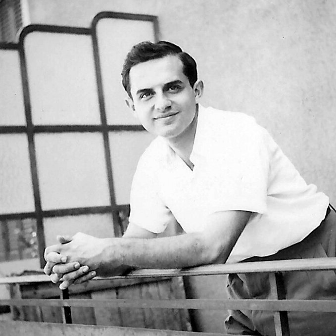 L'image montre un jeune homme posant avec un sourire amical. Il est vêtu d'une chemise blanche et de pantalons clairs, se tenant sur un balcon avec des barres métalliques. L'arrière-plan présente une architecture simple, avec des fenêtres et des volets. L'ambiance est décontractée et ensoleillée, suggérant un moment de détente.