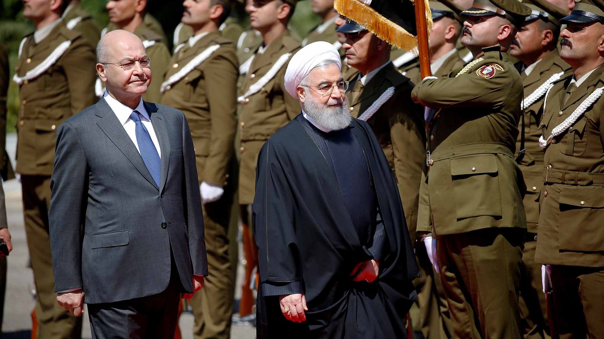 الصورة تظهر شخصيتين سياسيين يسيران معًا، حيث يبدو أنهما في مراسم رسمية. الشخص الأول يرتدي بدلة رسمية، بينما الشخص الثاني يرتدي زيًا تقليديًا أسود مع عمامة. خلفهم يظهر حرس الشرف يرتدون uniforms عسكرية، ويحملون الأعلام. الأجواء تعكس طابعًا رسميًا وجديًا، كما أن الإضاءة واضحة مما يدل على أنها تم التقاطها خلال النهار.