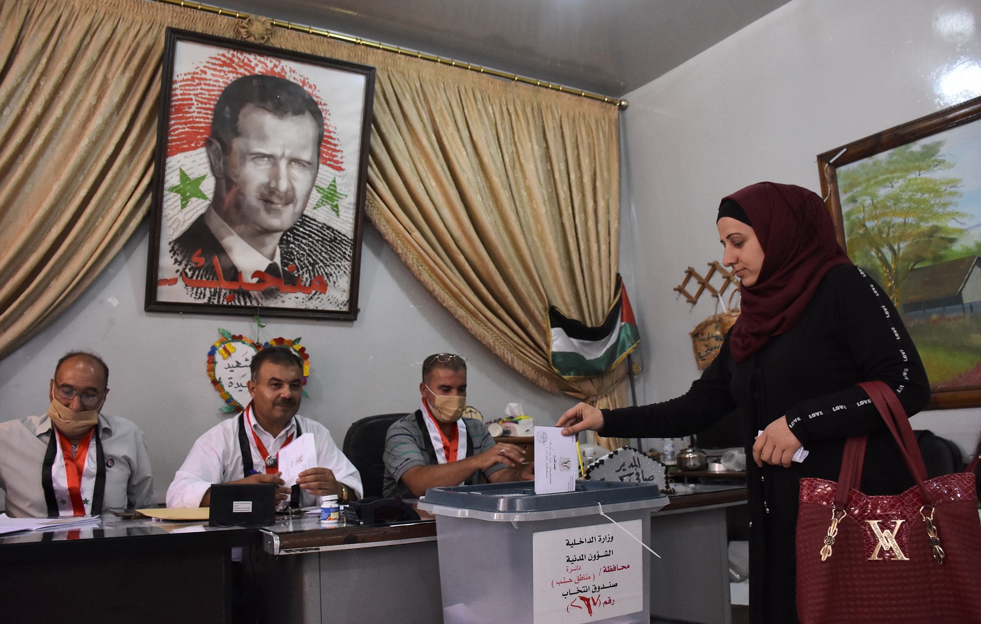 تظهر الصورة امرأة ترتدي عباءة وحجابًا تضع ورقة اقتراع في صندوق الانتخاب. خلفها، يوجد لوحة كبيرة تحمل صورة شخصية معروفة. يجلس ثلاثة رجال خلف طاولة، حيث يبدو عليهم الاستعداد لمراقبة العملية الانتخابية. المكان يبدو رسميًا، مع ستائر وخلفية تشير إلى الطابع المحلي.