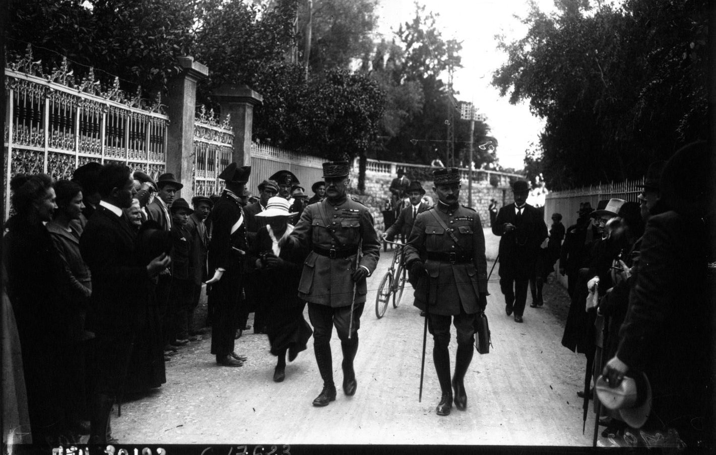 L'image montre une rue entourée de personnes, dont des militaires en uniforme. Deux officiers marchent au premier plan, semblant discuter. On aperçoit des civils sur les côtés, certains regardant vers les officiers. L'ambiance semble solennelle, et l'environnement est arboré, avec des bâtiments en arrière-plan. Les vêtements des personnes indiquent une époque historique. L'image a une qualité noire et blanche, typique des photographies anciennes.