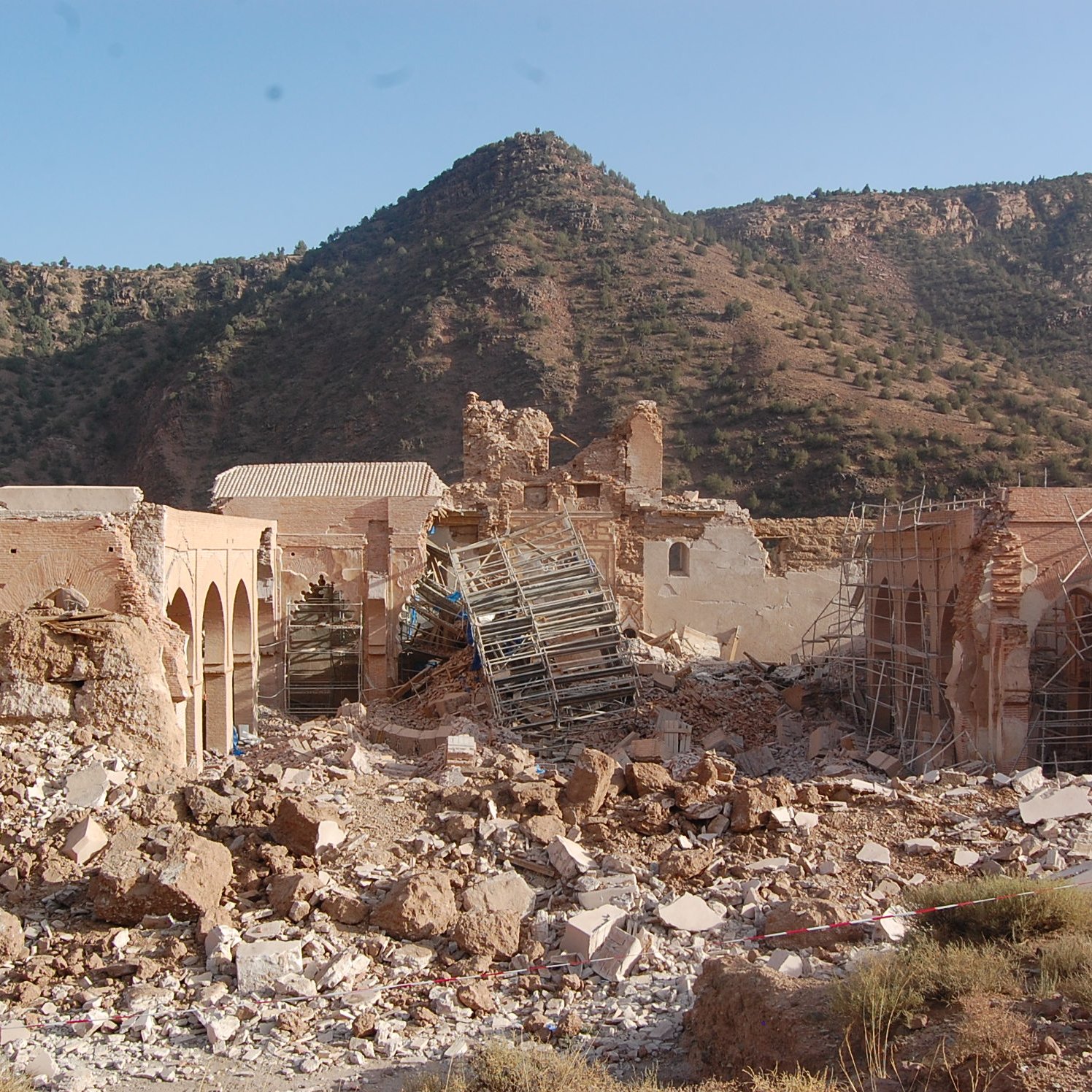 L'image montre un site de ruines où les structures sont partiellement effondrées, avec des débris éparpillés sur le sol. Des échafaudages sont visibles autour des bâtiments, suggérant un effort de restauration ou de démolition. En arrière-plan, on aperçoit une montagne aux pentes douces, recouverte de verdure, sous un ciel dégagé. L'ensemble dégage une impression de désolation mêlée à la beauté naturelle environnante.