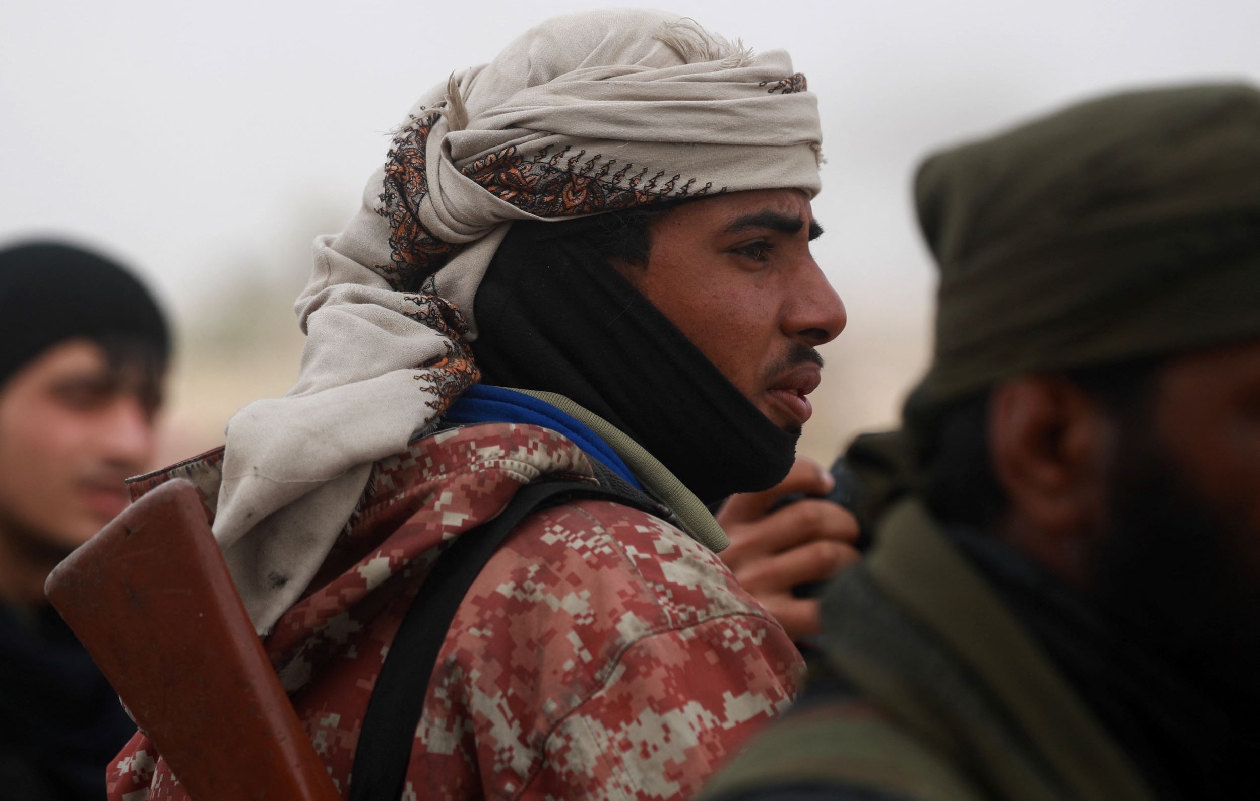 L'image montre un groupe d'individus, dont l'un est au premier plan. Il porte un couvre-chef traditionnel et une écharpe, ce qui lui donne un aspect distinctif. Son expression est sérieuse, et il semble concentré. Il tient également ce qui semble être une arme, indiquant un contexte potentiellement militaire ou de conflit. L'arrière-plan est flou, avec d'autres personnes visibles, suggérant une atmosphère de rassemblement ou d'affrontement.