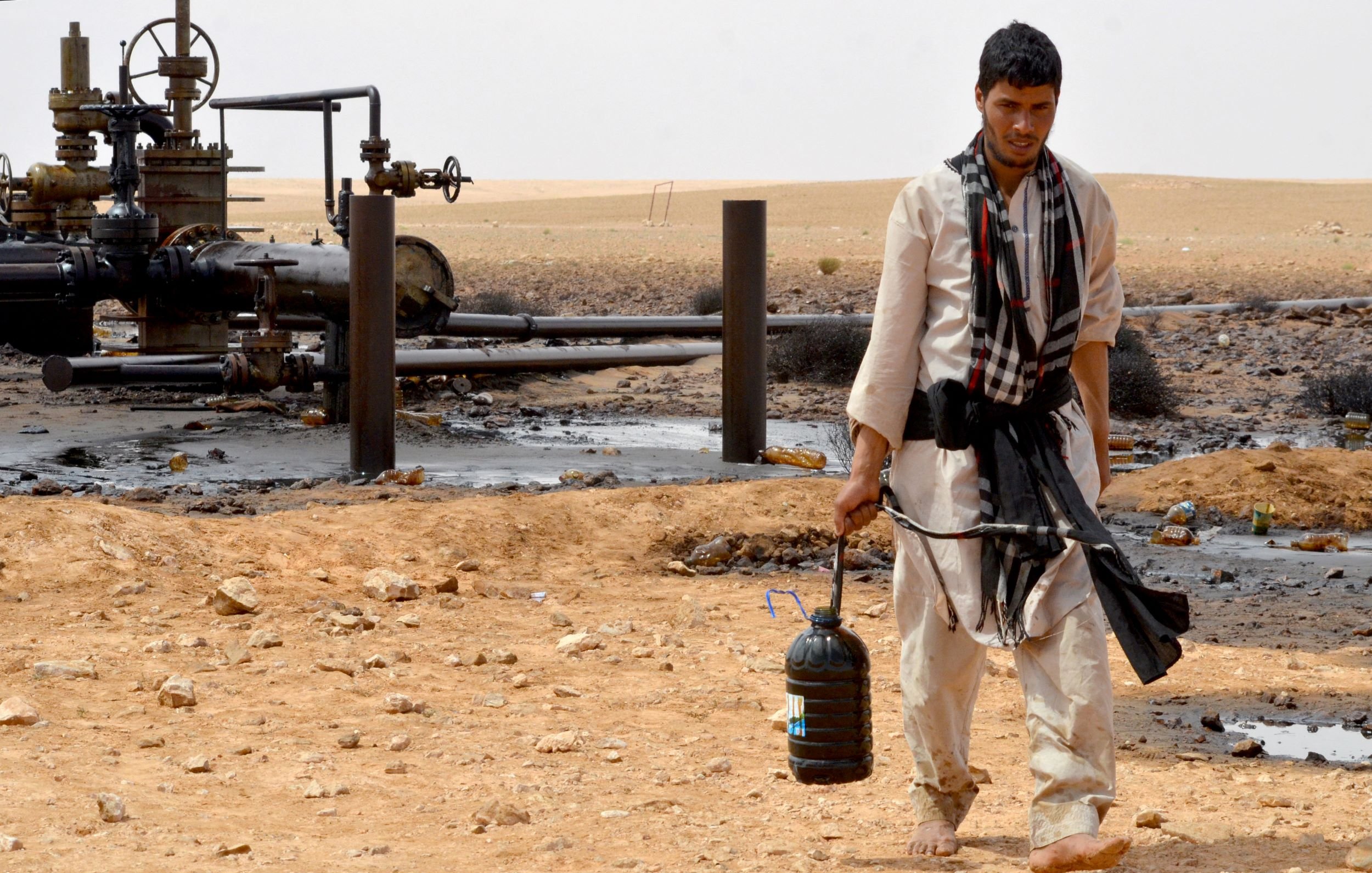 L'image montre un homme marchant à travers un paysage désertique. Il porte des vêtements traditionnels et a un seau noir dans une main. En arrière-plan, on peut voir des installations liées à l'industrie pétrolière, avec des tuyaux et des structures. Le sol est parsemé de pierres et il y a des traces de pétrole sur le terrain. L'environnement semble aride et désertique.