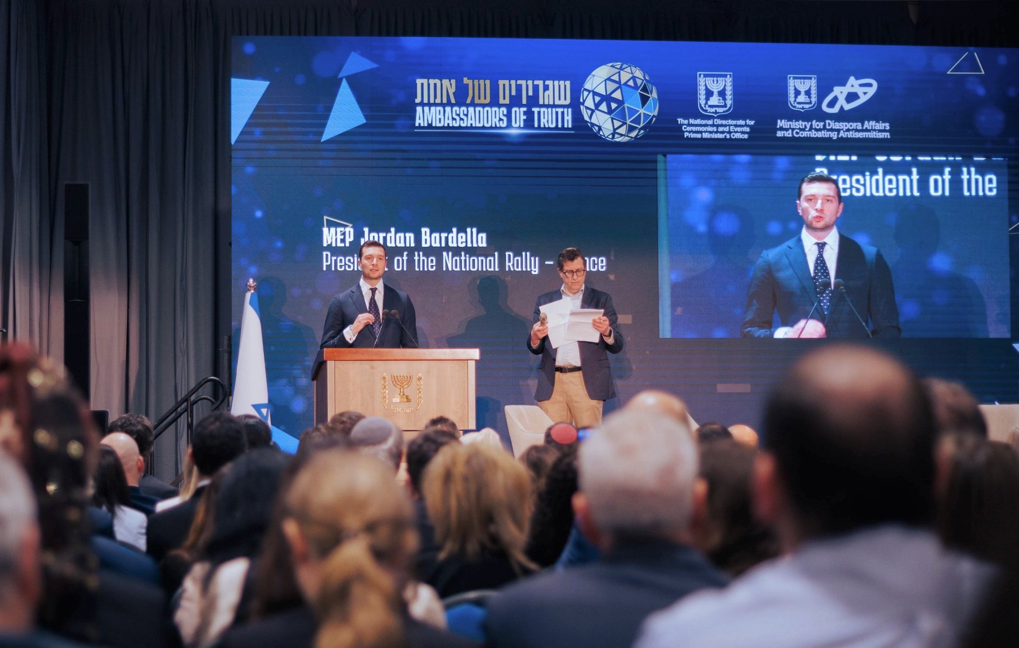 الصورة تظهر حدثًا رسميًا حيث يتحدث متحدث أمام جمهور كبير. على المنصة، يوجد شخص يحمل ورقة، ويبدو أن المتحدث يقرأ من نص. هناك خلفية تعرض شعارًا ومعلومات عن الفعالية. الأجواء تبدو جدية والر audience مهتم.