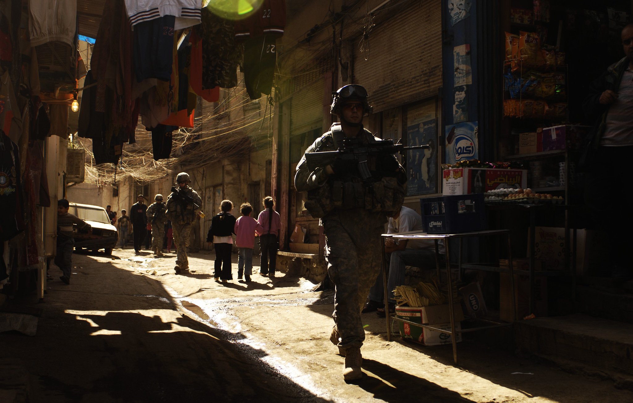 L'image montre une rue étroite et animée typique d'un marché urbain. Des soldats en uniforme, équipés d'armes, marchent le long de la ruelle. On aperçoit également des enfants qui semblent jouer ou observer les soldats. Les bâtiments sont proches les uns des autres, et des vêtements sont suspendus au-dessus de la rue. La lumière du soleil filtre à travers la ruelle, créant une ambiance contrastée entre l'ombre et la lumière. L'environnement semble à la fois vivant et chargé de tension.
