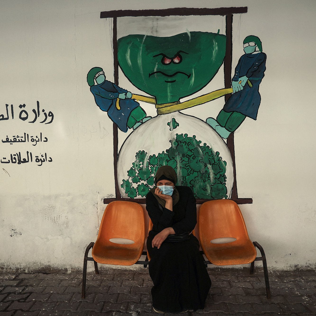 L'image montre une femme assise sur un banc orange, dans une ambiance urbaine. Elle porte un vêtement traditionnel noir et a un masque sur le visage. Derrière elle, on peut voir une fresque murale représentant deux personnages en tenue de protection, qui semblent tirer sur une grande horloge en forme de verre à sablier contenant du matériel de santé, symbolisant probablement une thématique liée à la santé publique. Le texte en arabe indique "Ministère de la Santé". L'ensemble de la scène évoque des questions de santé et peut-être de vigilance face à des défis sanitaires.