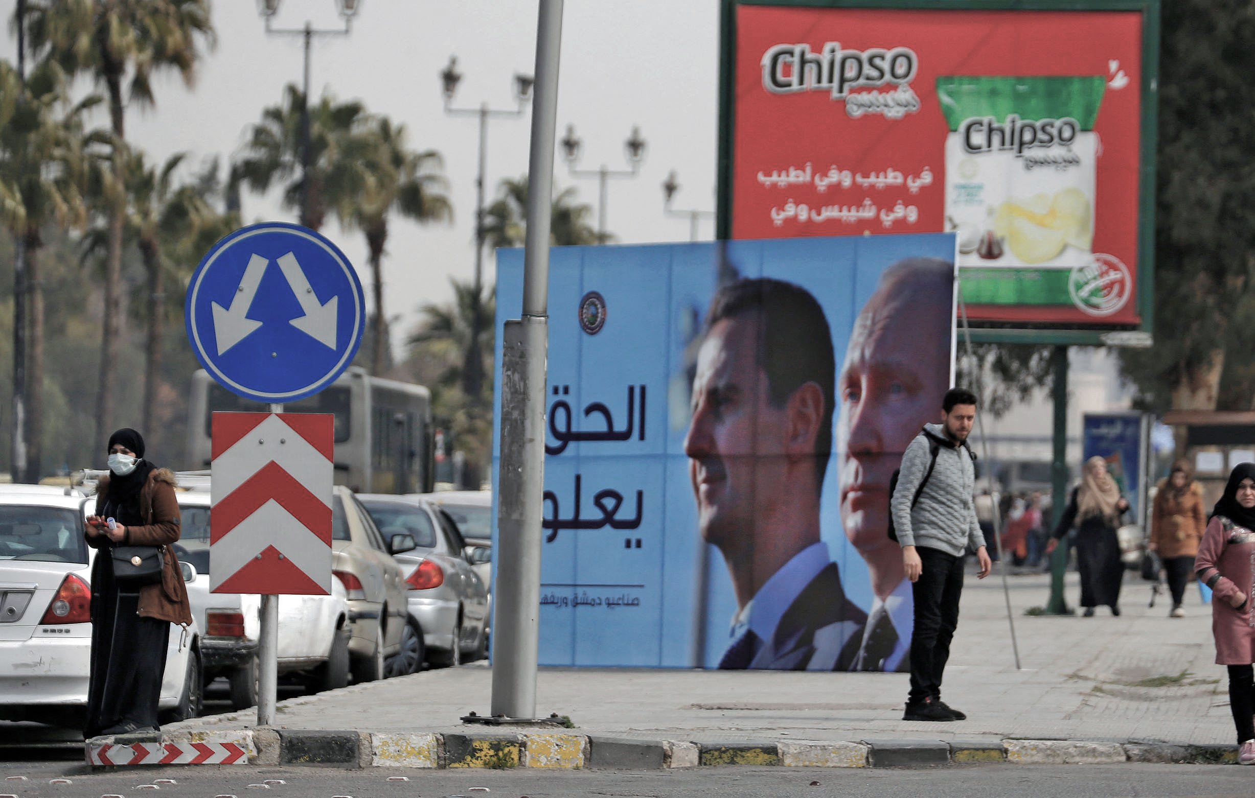 در این تصویر، خیابانی مطرح شده و در آن دو نفر در کنار پیاده‌رو ایستاده‌اند. در پس‌زمینه، چندین خودرو پارک شده و همچنین یک بیلبورد بزرگ با تصاویر و متنی به زبان عربی دیده می‌شود. این بیلبورد به احتمال زیاد به مقامات و موضوعات سیاسی اشاره دارد. تبلیغاتی برای یک برند معروف تنقلات نیز در بالای تصویر دیده می‌شود. در مجموع، تصویر حال و هوای شهری را به نمایش می‌گذارد.