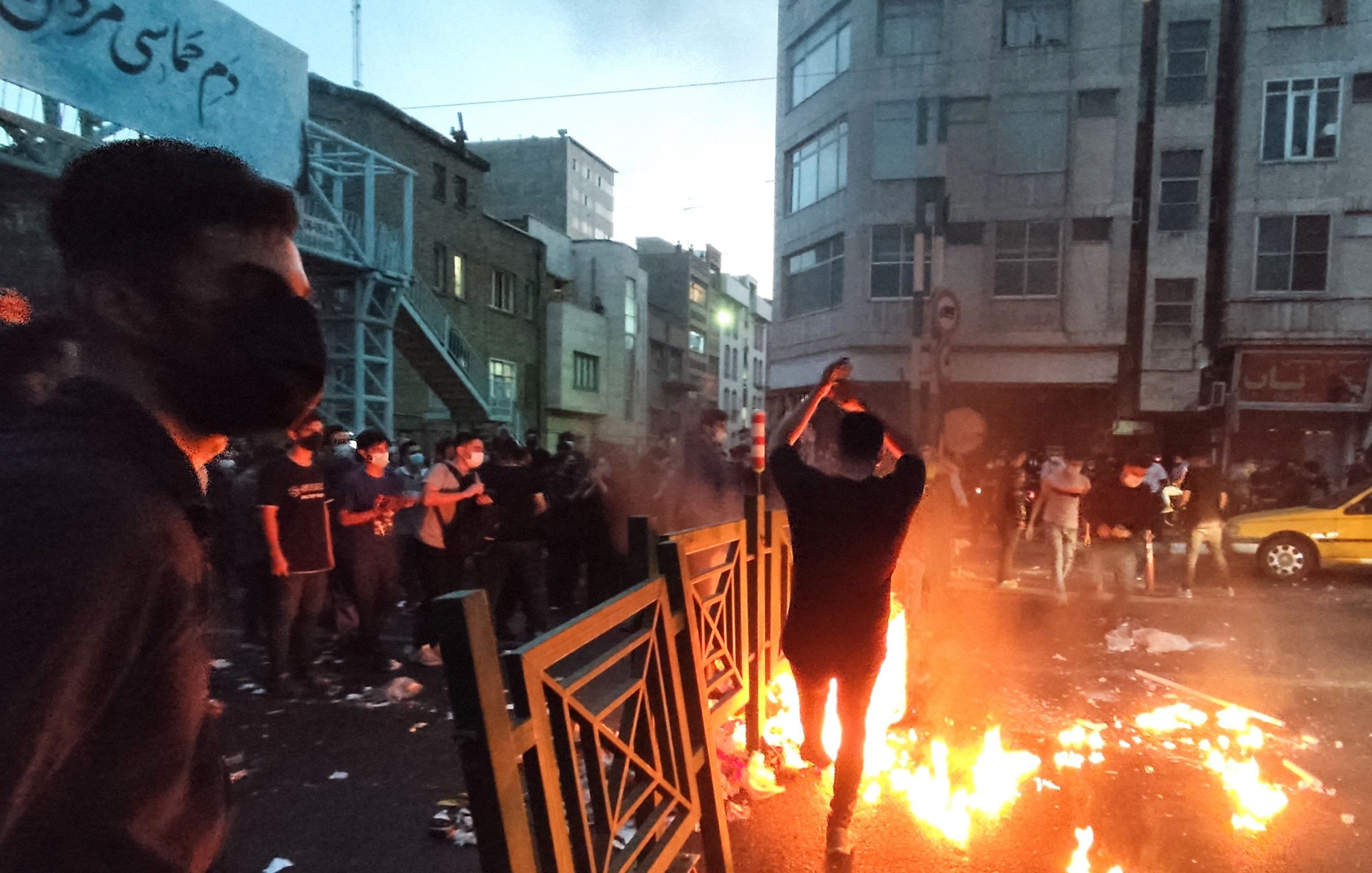 در این تصویر، گروهی از افراد در خیابانی در حال تظاهرات هستند. در مرکز عکس، فردی در حال آتش زدن شیئی است که آتش به‌طور واضحی شعله‌ور شده است. دور تا دور او زباله‌ها و آشغال‌ها پراکنده شده و برخی از معترضان در حال تماشا هستند. بیشتر افراد ماسک به‌دهن دارند و فضای عمومی به نظر شلوغ و تنش‌زا می‌رسد. ساختمان‌های اطراف و نور پایین آسمان نیز به حالت دراماتیک تصویر افزوده است.