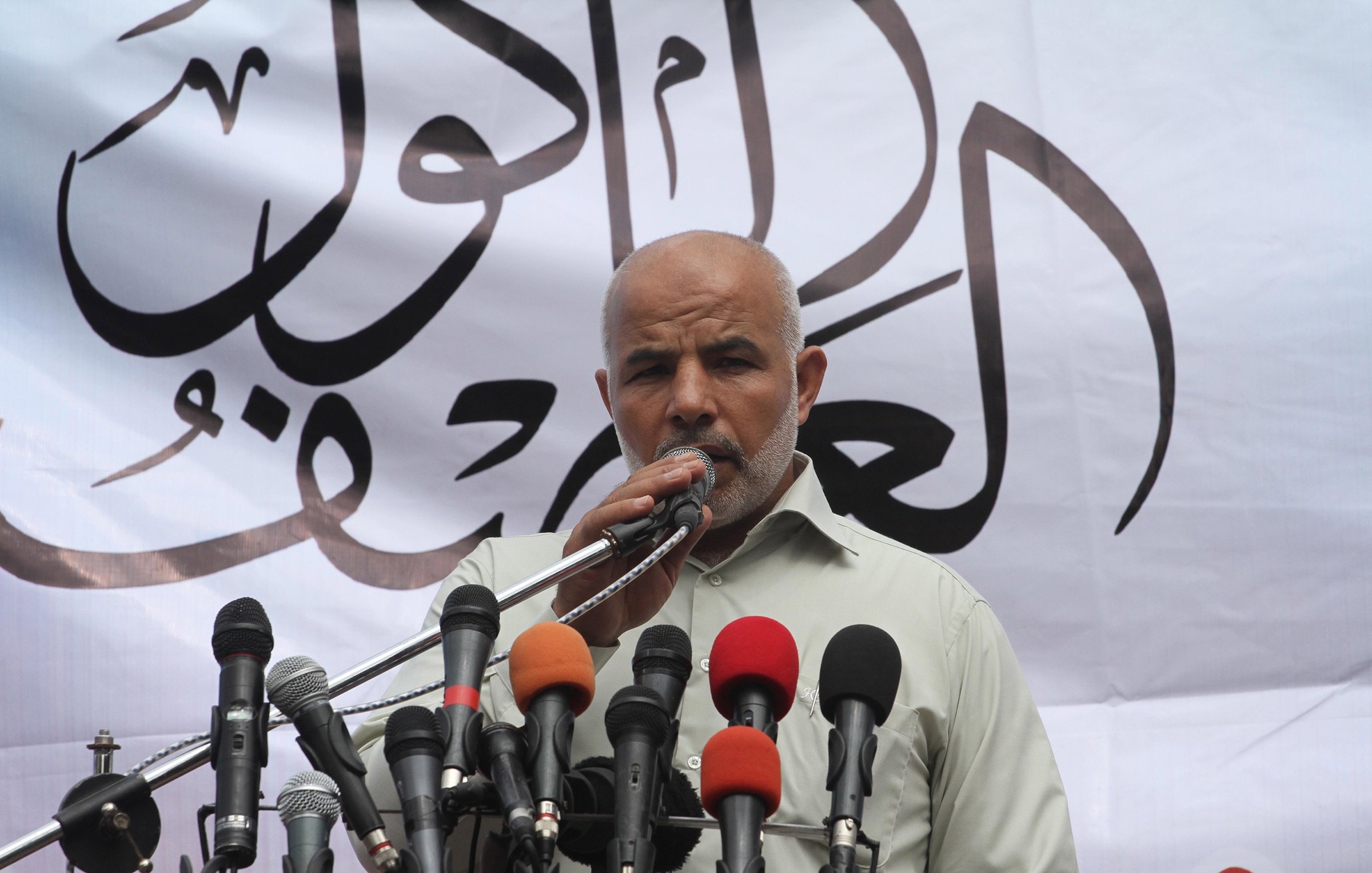 L'image montre un homme debout devant un microphone, prenant la parole lors d'un événement. Il a une barbe grise et porte une chemise claire. Derrière lui, un grand panneau blanc est décoré d'écritures arabes. Sur le devant, plusieurs microphones de différentes couleurs sont alignés, ce qui indique qu'il s'agit probablement d'une conférence de presse ou d'un discours public. L'atmosphère semble solennelle et engagée.