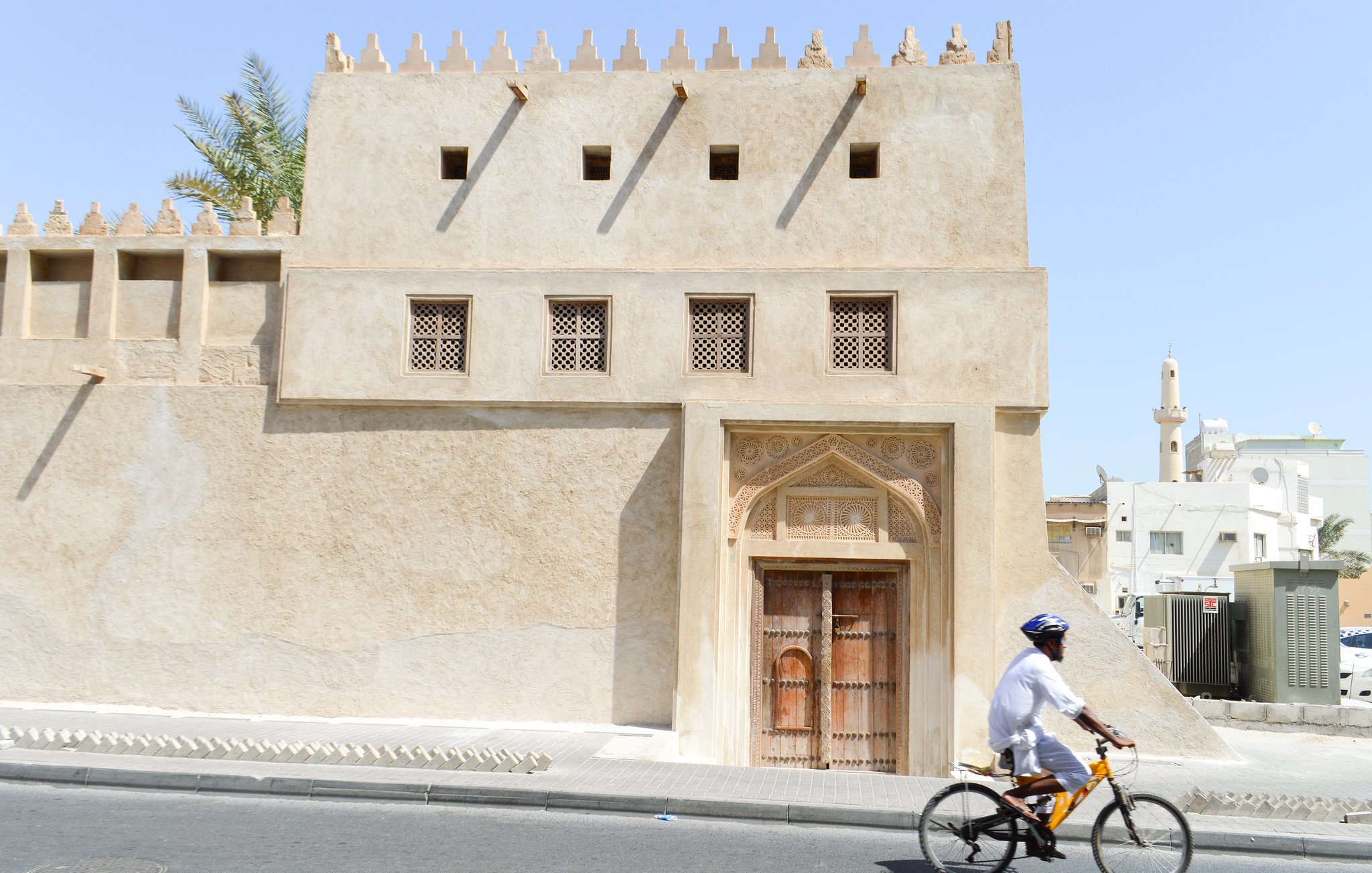 La imagen muestra un edificio de arquitectura tradicional, con paredes de color beige y detalles ornamentales en la fachada. La puerta principal es de madera tallada, y hay ventanas con rejillas decorativas. Al lado de la construcción, un ciclista vestido con ropa ligera y un casco pedalea por una calle pavimentada. El ambiente parece cálido, lo que sugiere un clima árido, típico de ciertas regiones. En el fondo, se pueden observar otros edificios y estructuras, que contribuyen a la sensación de un entorno urbano.