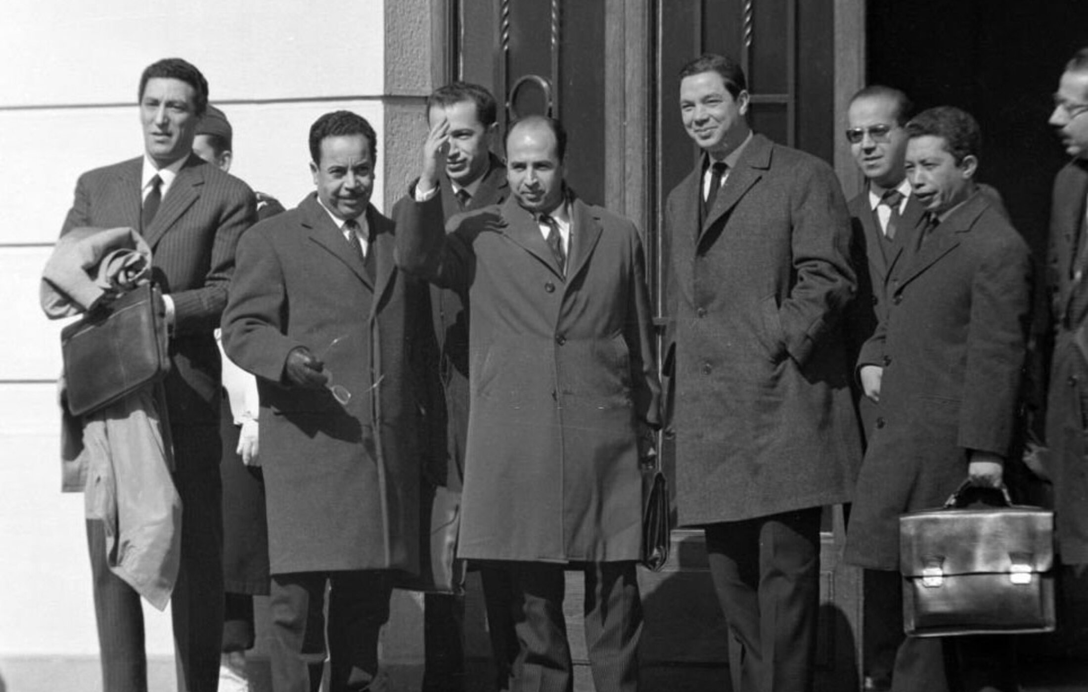 L'image montre un groupe de plusieurs hommes en costume, se tenant à l'extérieur d'un bâtiment. Ils portent des manteaux d'hiver, indiquant que la scène se déroule probablement par temps frais. L'un des hommes, au centre, semble être en train de saluer ou de parler avec enthousiasme. Les autres hommes affichent des expressions neutres ou souriantes. L'environnement est urbain, avec des détails architecturaux en arrière-plan.