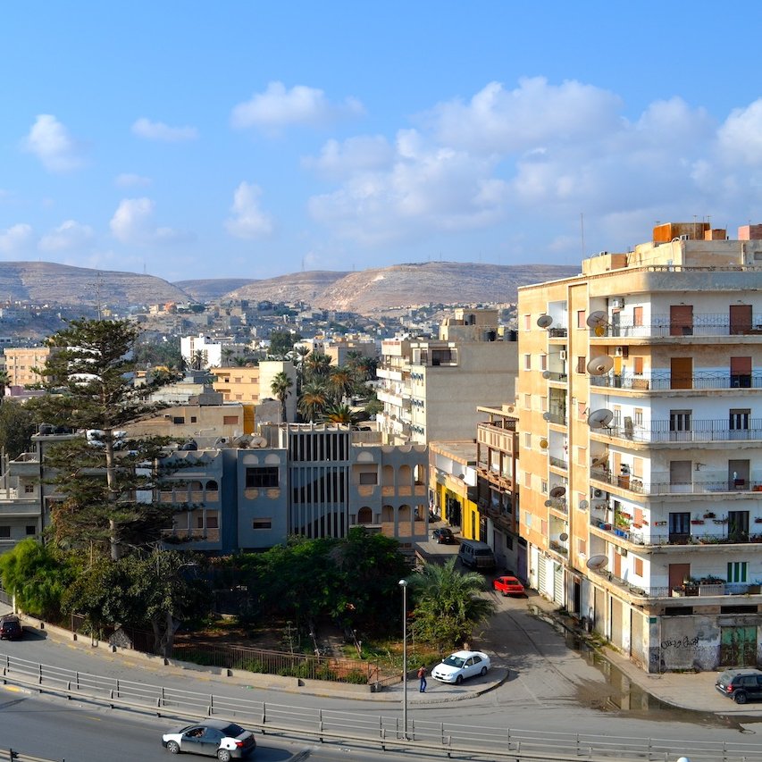 L'image montre un paysage urbain avec des bâtiments variés, y compris des immeubles résidentiels et commerciaux, ainsi qu'une route serpentine. Au loin, on peut voir des collines et un ciel clair avec quelques nuages. La scène dégage une ambiance paisible, avec des voitures circulant sur la route et des arbres ajoutant une touche de verdure à l'environnement.