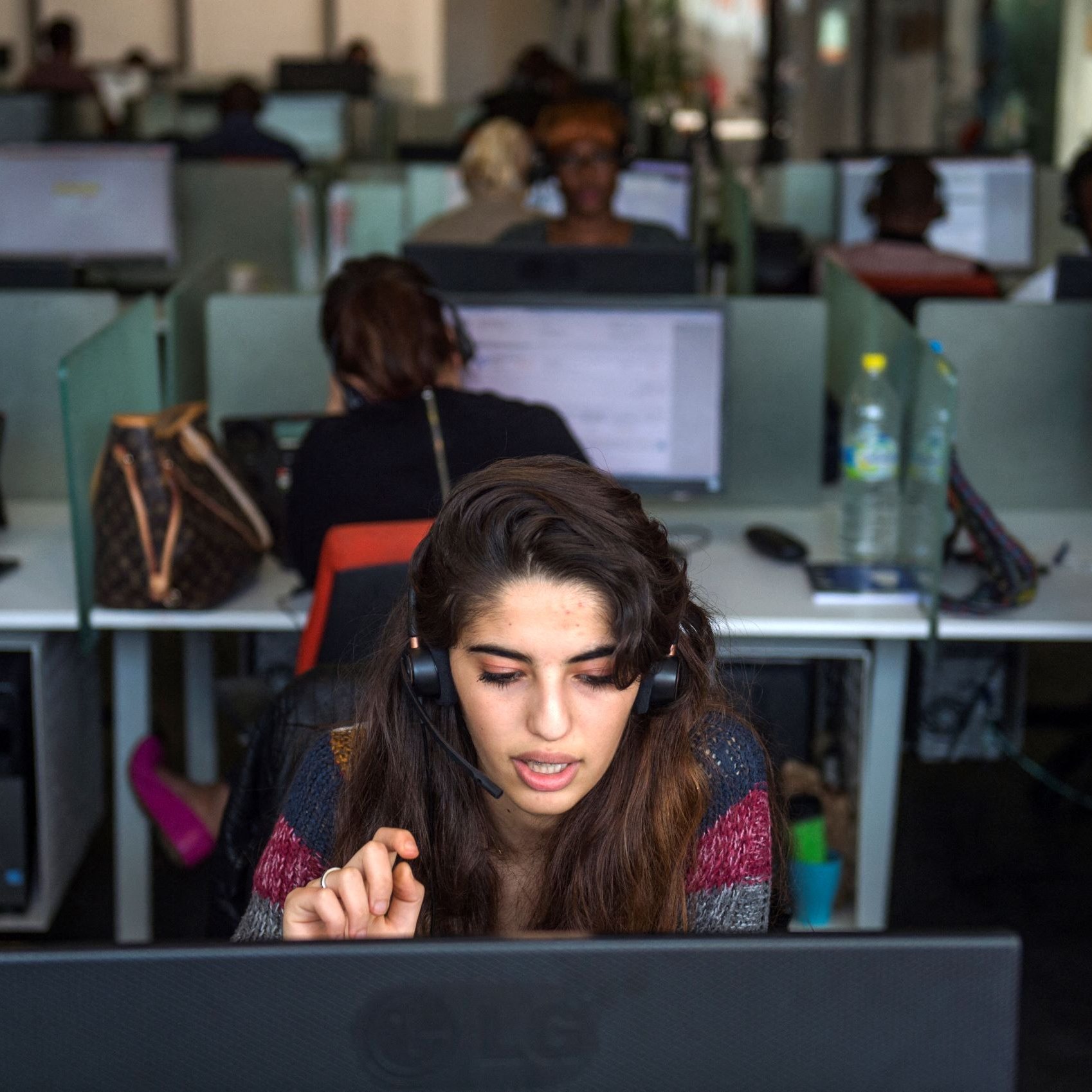 The image depicts a call center environment. In the foreground, a young woman is wearing a headset, concentrating intently on her work at a computer. She appears to be engaged in a conversation, with one hand raised, possibly indicating she is speaking or asking a question. In the background, several other people are working at similar desk setups, each using computers and headsets, which creates a bustling atmosphere. The workspace is well-lit and modern, with a mix of personal items placed at some desks.