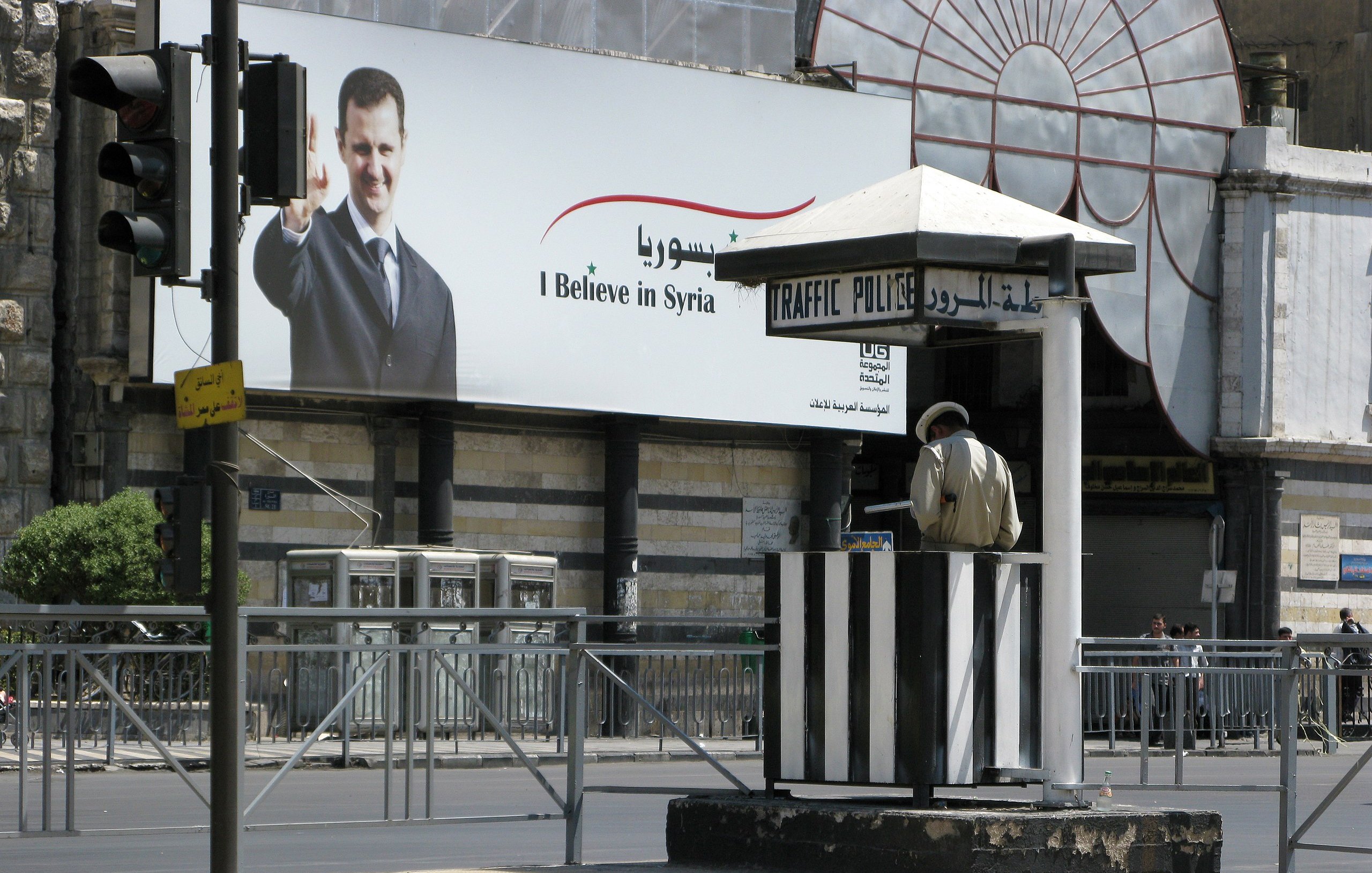 L'immagine mostra una cabina di polizia del traffico situata in una strada. In secondo piano, c'è un grande manifesto che ritrae un uomo che saluta, con il testo in arabo e italiano che dice "I Believe in Syria". La scena è animata ma appare tranquilla, con poche auto visibili e un'illuminazione chiara. Il contrasto tra la cabina di polizia e il manifesto può suggerire elementi di sicurezza e comunicazione politica.