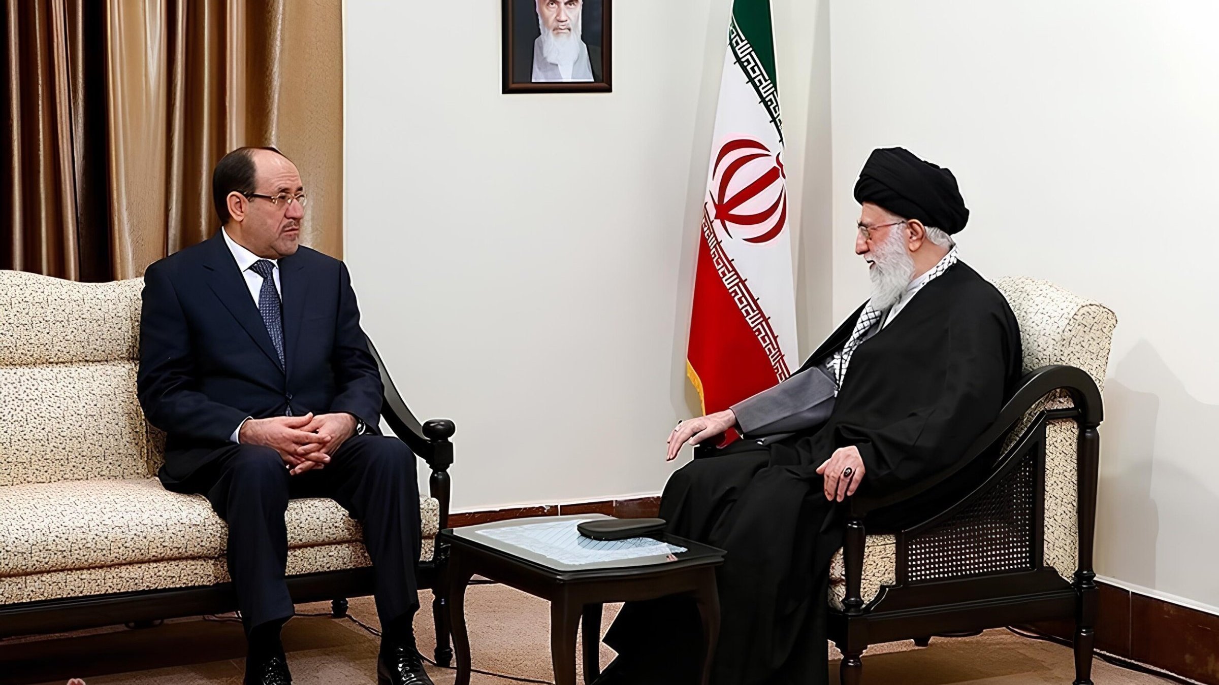 Due uomini seduti in una sala con bandiera iraniana, discutono seriamente.