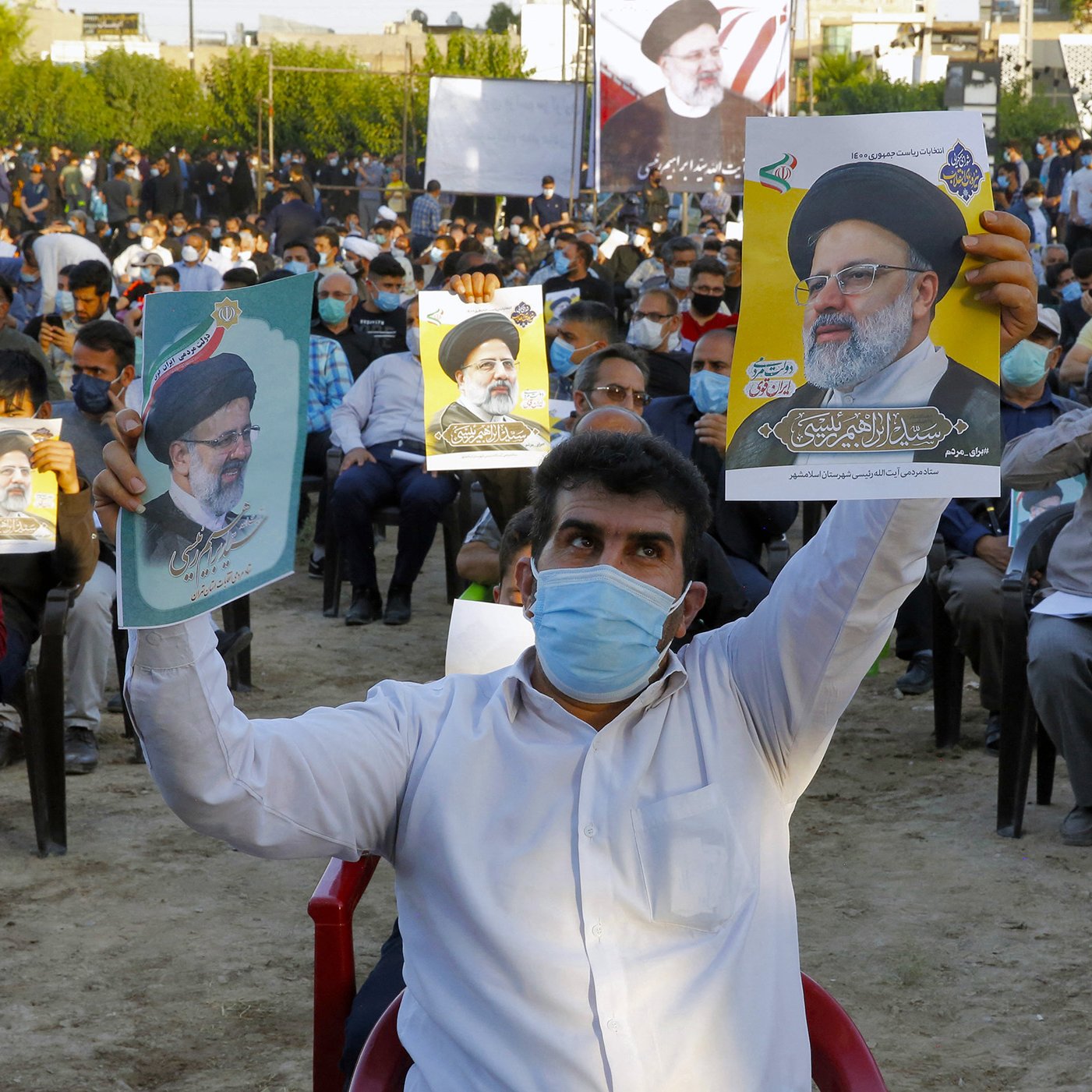 L'image montre une assemblée de personnes rassemblées en plein air, assises sur des chaises. La plupart des participants portent des masques. Un homme, au premier plan, lève une affiche représentant une figure politique, probablement un leader ou un candidat, symbolisant un soutien. Les autres personnes autour de lui tiennent également des affiches similaires. En arrière-plan, on peut apercevoir des arbres et des structures, suggérant un rassemblement organisé. L'atmosphère semble être celle d'un événement politique ou d'une manifestation.