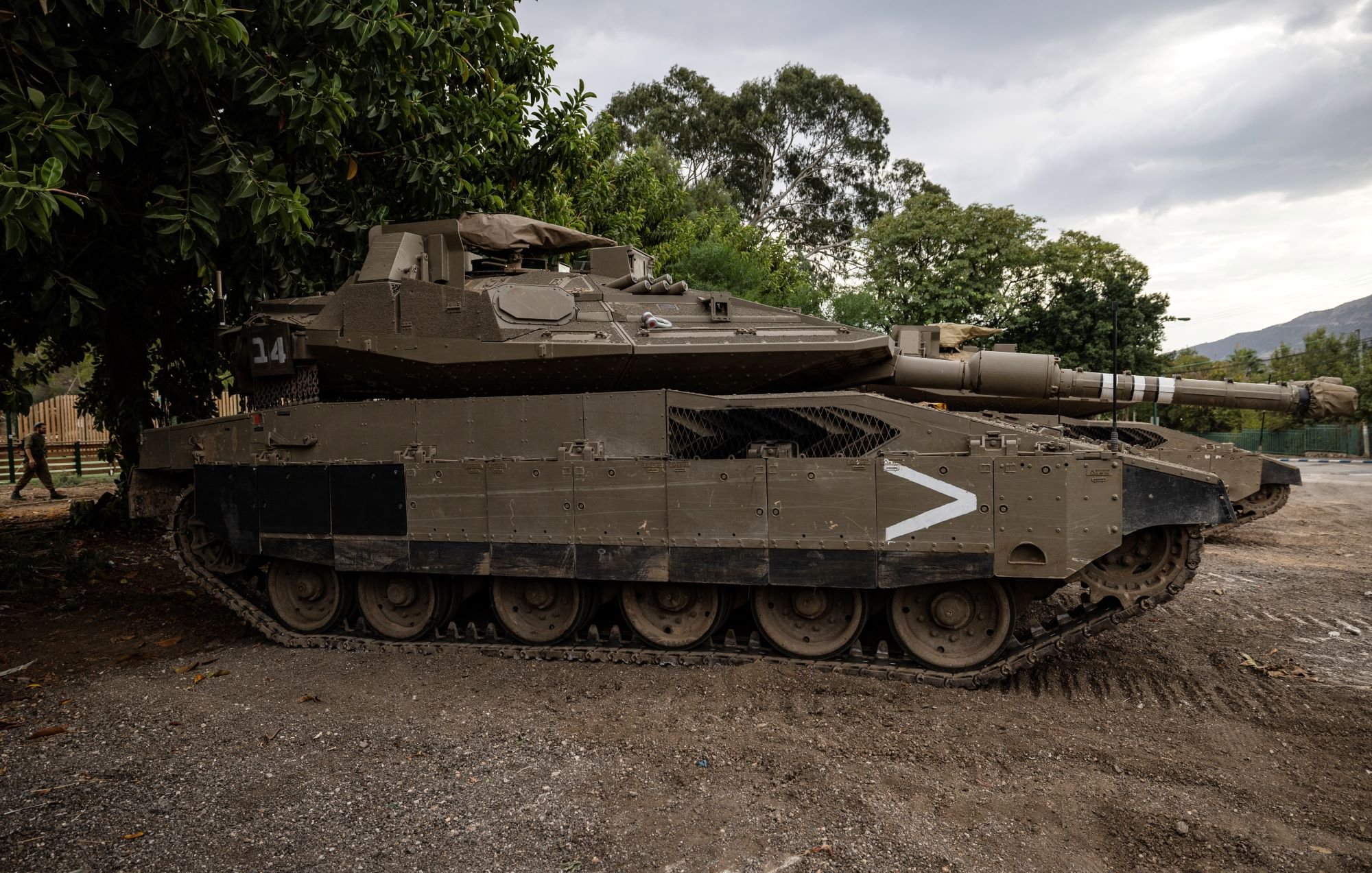 L'image montre un char d'assaut stationné sur un terrain. Le véhicule est de couleur verte foncée et possède des marques de camouflages. Il est équipé d'une tourelle avec un canon pointé vers l'avant. En arrière-plan, on peut voir des arbres et un ciel nuageux, ce qui donne une ambiance militaire à la scène. Le char semble robuste et bien conservé, prêt pour une utilisation éventuelle.