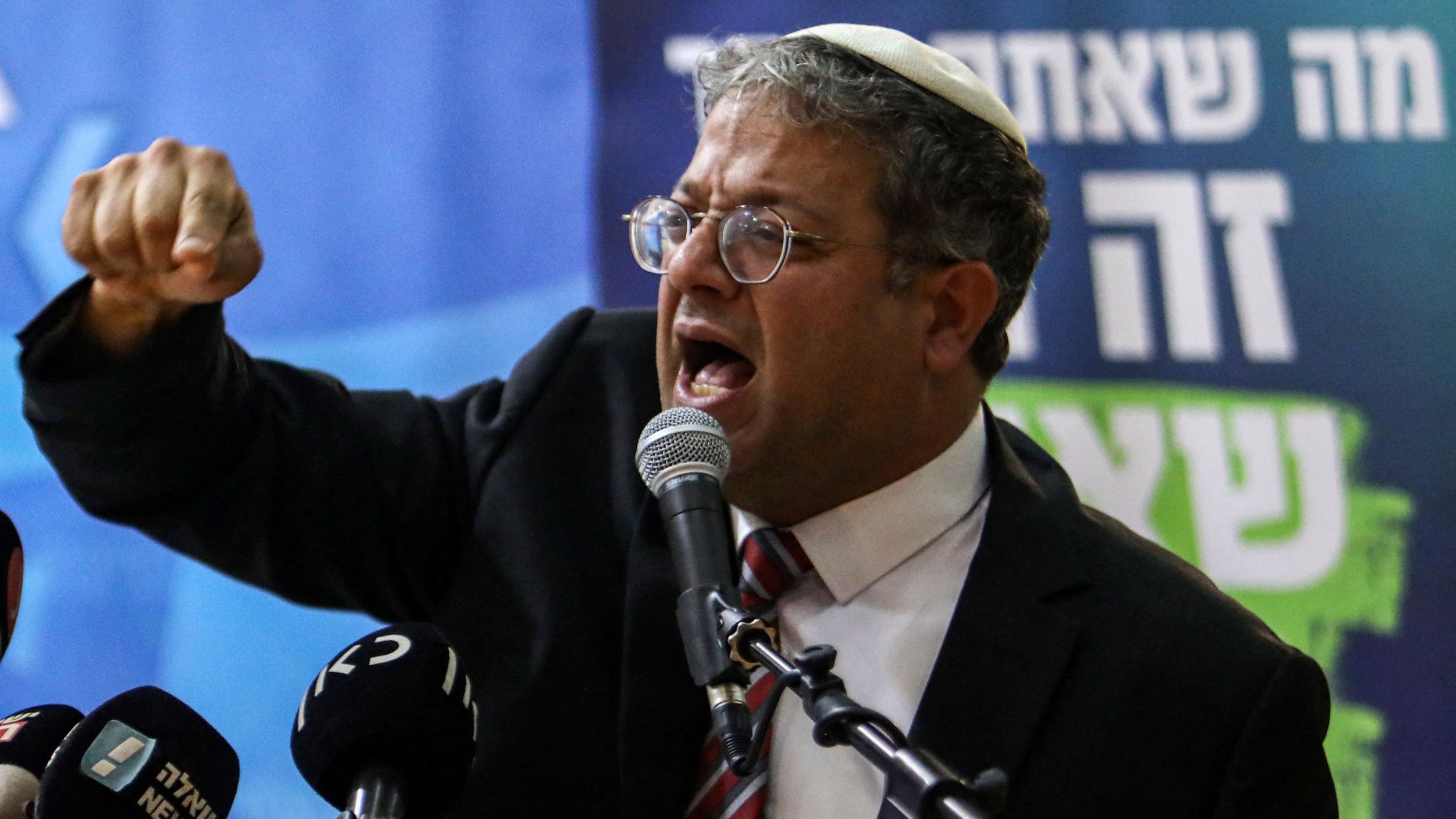 Nell'immagine si vede un uomo che parla appassionatamente al microfono, indossando un kippah. Sembra un politico o un oratore, con espressione intensa e gesti enfatici mentre esprime le sue idee. Sullo sfondo ci sono delle bandiere e scritte che potrebbero indicare un evento politico. L'atmosfera appare energica e coinvolgente.