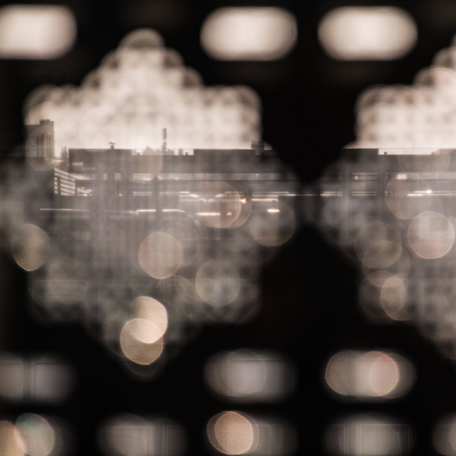 L'image montre une vue floue à travers une structure découpée, de formes géométriques. On peut apercevoir des silhouettes de bâtiments en arrière-plan, avec une lumière réfléchie qui crée des éclats. L'atmosphère générale semble douce et légèrement mystique, accentuée par les jeux de lumière et de flou. Les détails précis des éléments au loin ne sont pas nettement visibles, ce qui ajoute à l'effet artistique de la composition.