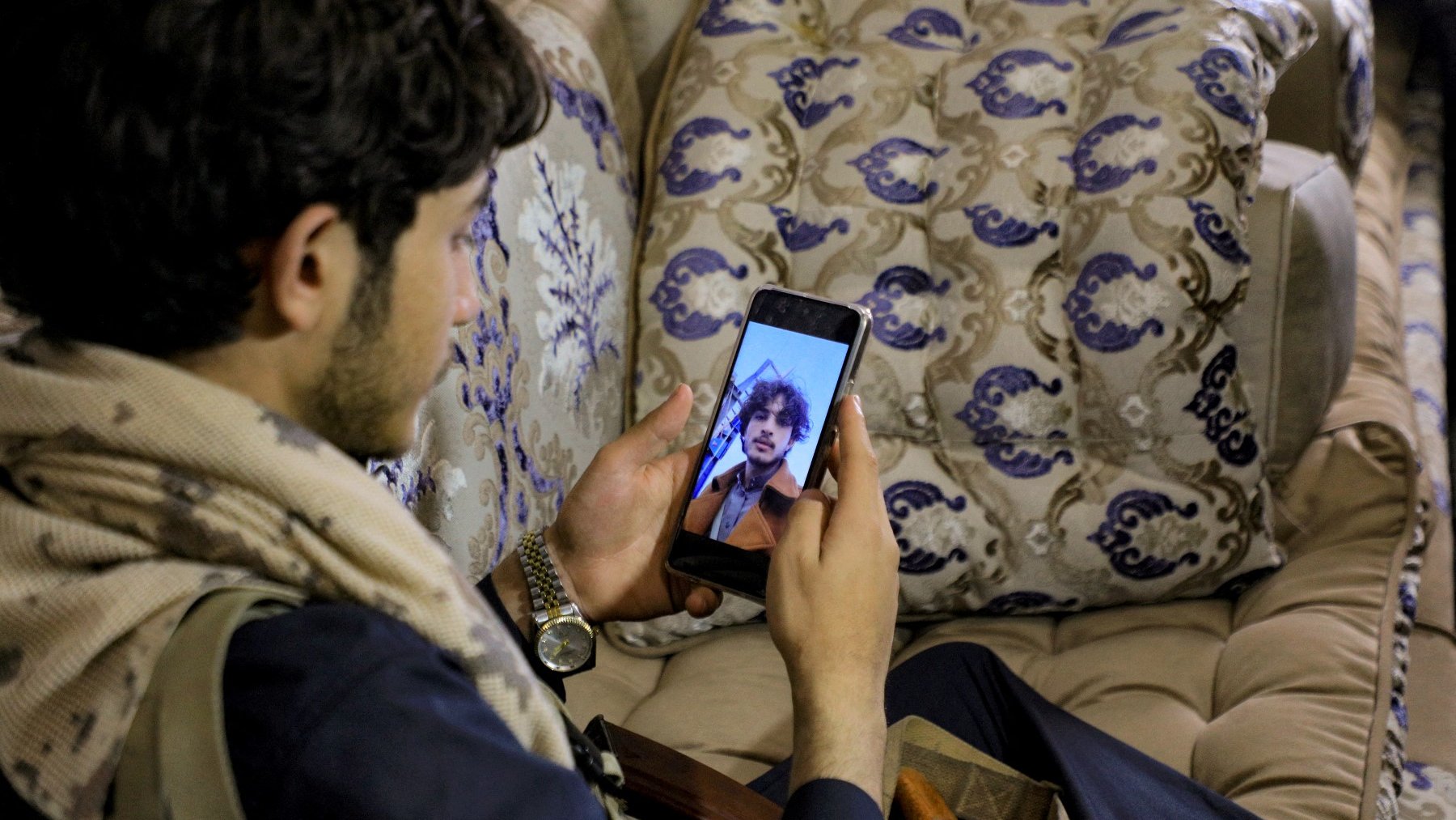 L'image montre un jeune homme assis sur un canapé décoré, regardant son téléphone portable. Il porte une écharpe et semble contemplatif. À côté de lui, on peut apercevoir une boîte et un objet, peut-être une arme ou un accessoire. Le décor est chaleureux et traditionnel, avec des coussins ornés de motifs élégants.