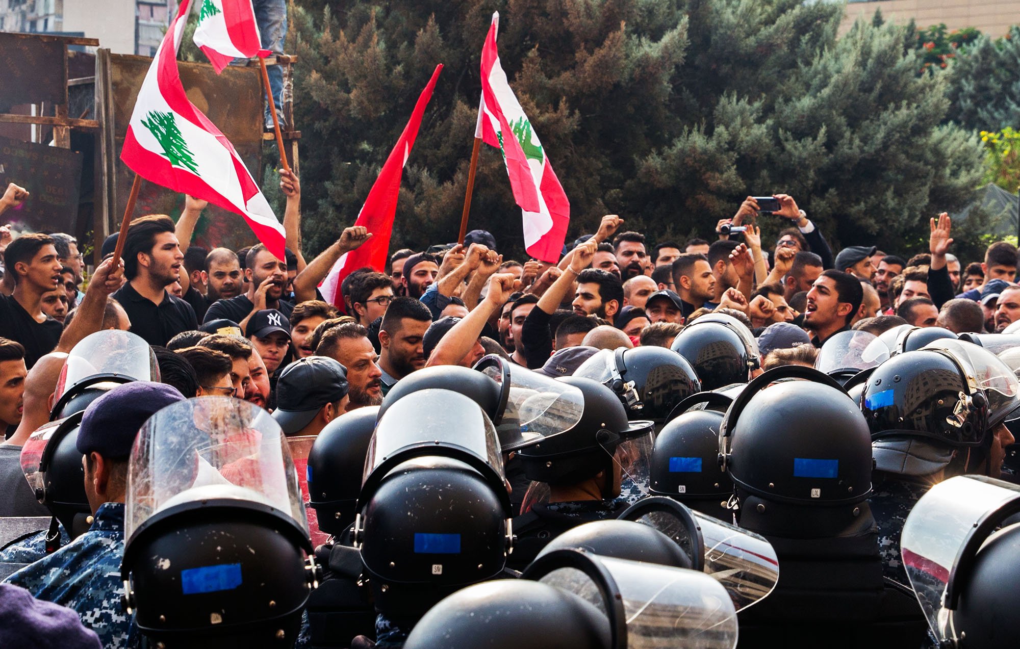 L'image montre une scène de manifestation avec un grand nombre de personnes brandissant des drapeaux libanais. Les manifestants semblent exprimer leurs revendications avec des poings levés, tandis qu'une ligne de policiers en uniforme, équipés de casques et de protections, se tient face à eux. L'ambiance est tense, illustrée par la proximité des manifestants et des forces de l'ordre. Des arbres en arrière-plan complètent le décor de cette confrontation.