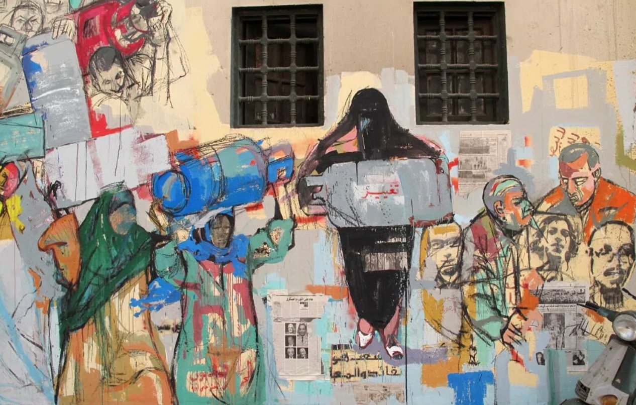 L'image montre une fresque murale colorée qui illustre diverses figures humaines engagées dans des activités sociales et politiques. Les personnages semblent porter des objets, portant des expressions variées, ce qui évoque des thèmes de lutte et de solidarité. Le mur est également décoré de collage de papiers et de textes, ajoutant une dimension visuelle riche et narrative à l'œuvre. Les couleurs utilisées sont vibrantes et contrastées, ce qui attire l'attention sur les détails et les émotions des personnages représentés.