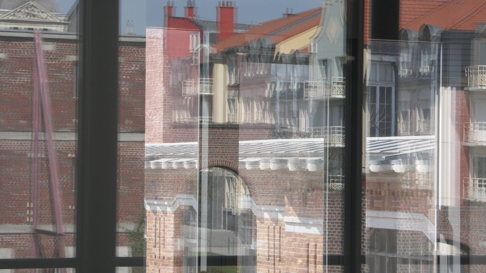 L'image montre un jeu de reflets à travers de grandes fenêtres. On peut apercevoir des bâtiments aux façades en briques et des toits de différentes couleurs. Les reflets créent une composition visuelle intrigante, mêlant les éléments extérieurs et la transparence des vitres. La lumière naturelle accentue les détails des structures architecturales.