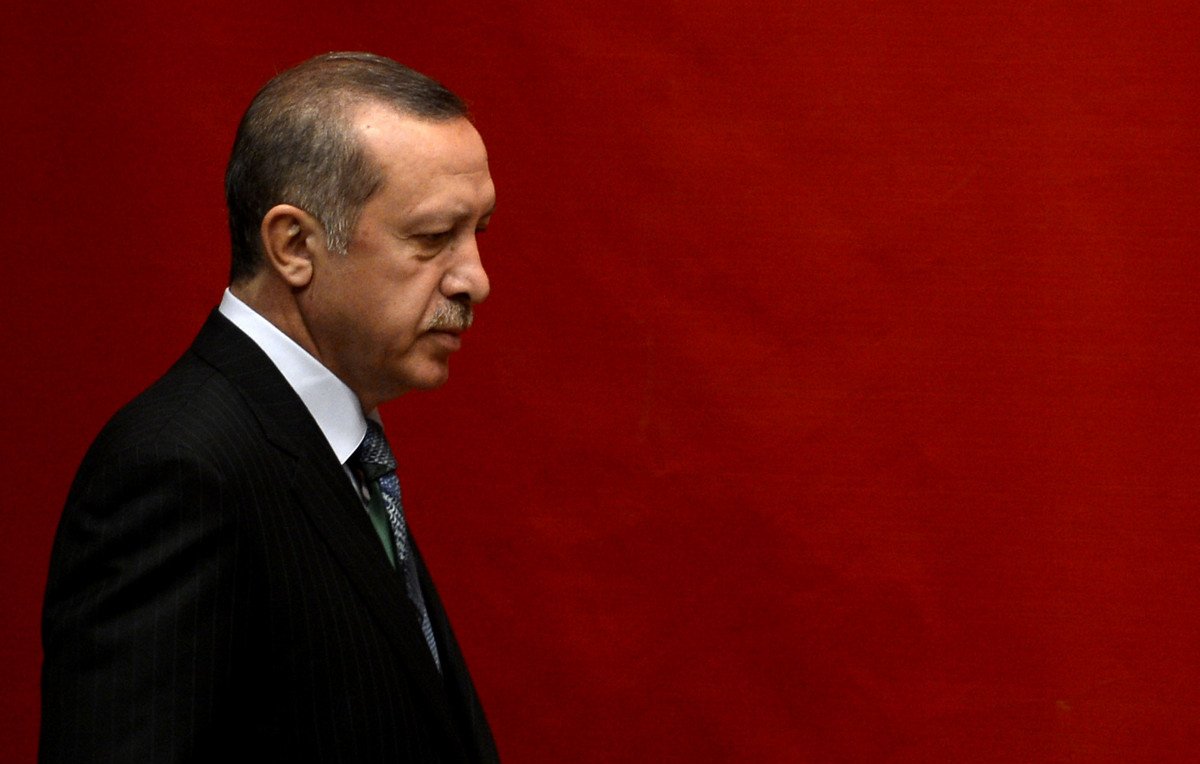 L'image montre un homme en costume sombre, marchant sur le côté, avec une expression sérieuse. Le fond est rouge uni, créant un contraste marqué avec sa tenue. L'éclairage met en avant son profil et accentue les détails de son visage.