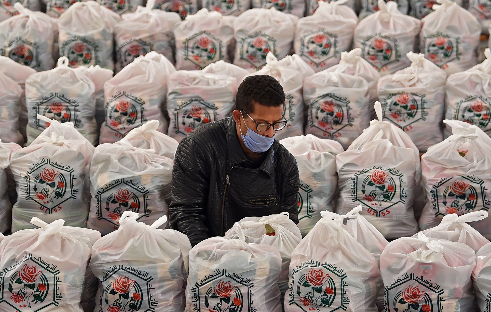 L'image montre un homme en train de travailler au milieu d'un grand espace rempli de sacs en plastique. Chaque sac est soigneusement empilé, arborant un motif de rose et des inscriptions en arabe. L'homme porte un masque et semble concentré sur sa tâche, probablement en train de trier ou de préparer quelque chose à l'intérieur des sacs. L'environnement est bien organisé, et la scène évoque un sentiment d'efficacité dans un contexte d'entraide ou de distribution.