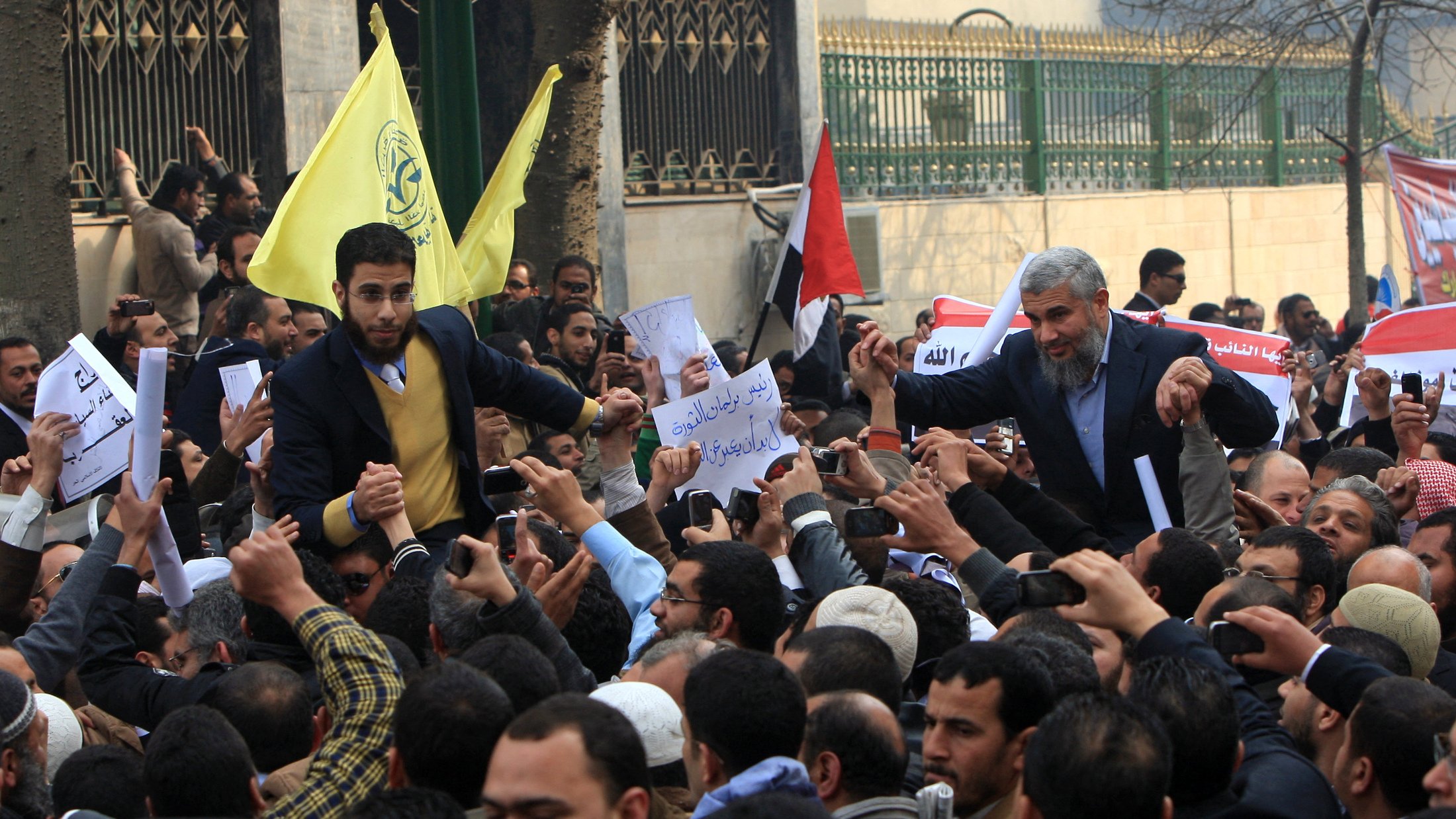 L'image montre une grande foule de personnes rassemblées, probablement lors d'une manifestation ou d'un rassemblement politique. On peut voir des individus tenant des pancartes et des drapeaux, exprimant des opinions ou des demandes. Les expressions des personnes sont variées, allant de l'enthousiasme à la détermination. L'ambiance semble énergique, reflétant un engagement collectif pour une cause. Les drapeaux et les signatures sur les pancartes suggèrent un contexte national ou politique spécifique.