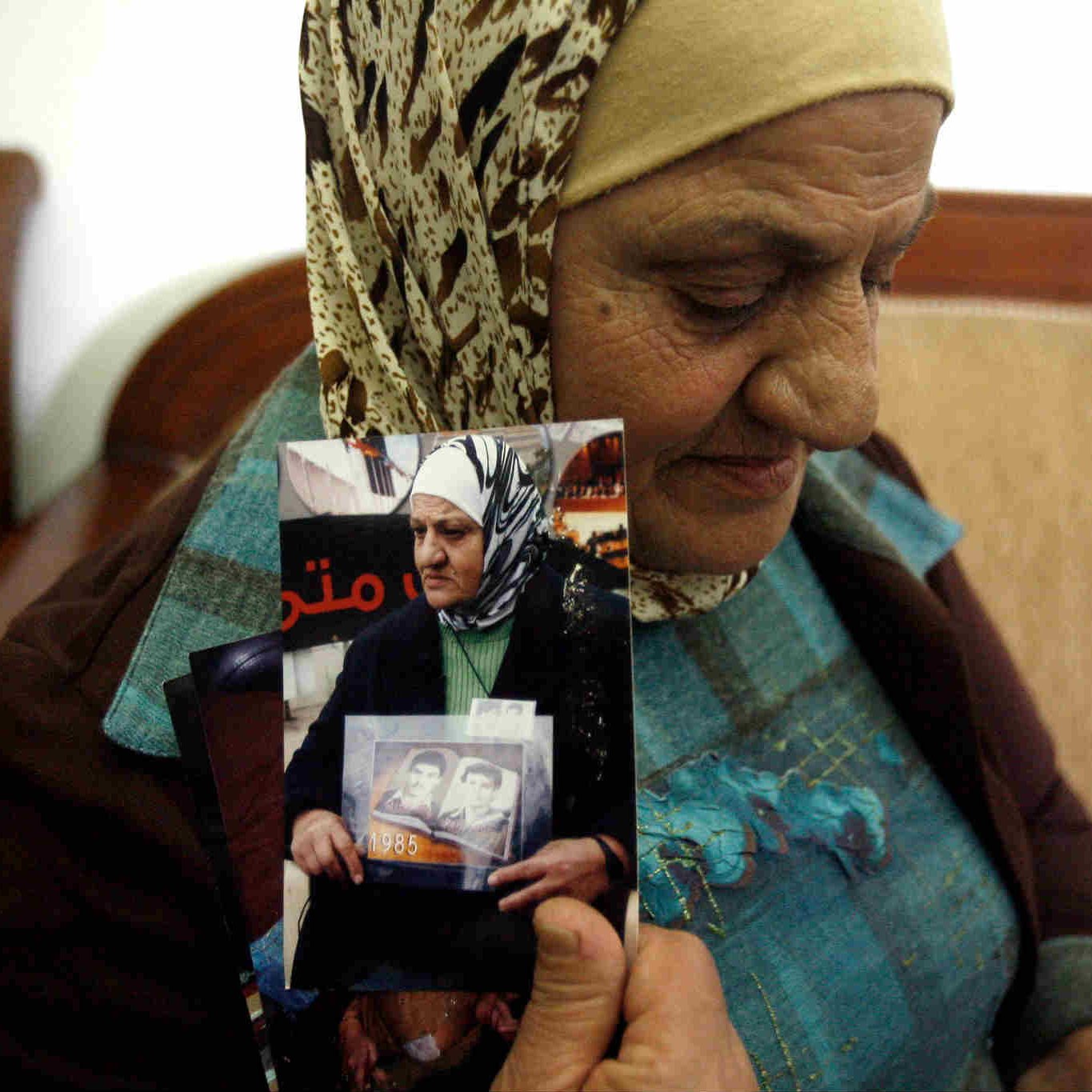 الصورة تظهر سيدة كبيرة في السن ترتدي حجابًا وتجلس على أريكة. تعبر ملامح وجهها عن التأمل والحنين، حيث تُمسك بعدد من الصور الفوتوغرافية في يديها. يبدو أن هذه الصور تحمل ذكريات مهمة بالنسبة لها، وقد تكون تتعلق بأفراد من عائلتها. الخلفية بسيطة وتُشير إلى بيئة منزلية مريحة.