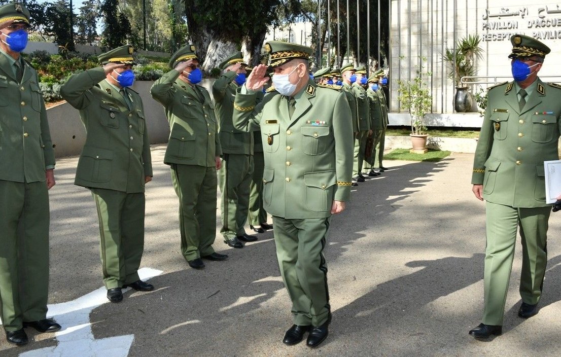 تظهر الصورة مجموعة من الضباط العسكريين يرتدون uniforms عسكرية باللون الأخضر، مع قبعات وأقنعة وجه زرقاء. يقوم الضباط بتحية أحد كبار الضباط الذي يسير في المقدمة. في الخلفية، يمكن رؤية مبنى حكومي أو عسكري مع بعض الأشجار والنباتات. يبدو أن اللحظة تشير إلى حدث رسمي أو استقبال عسكري.