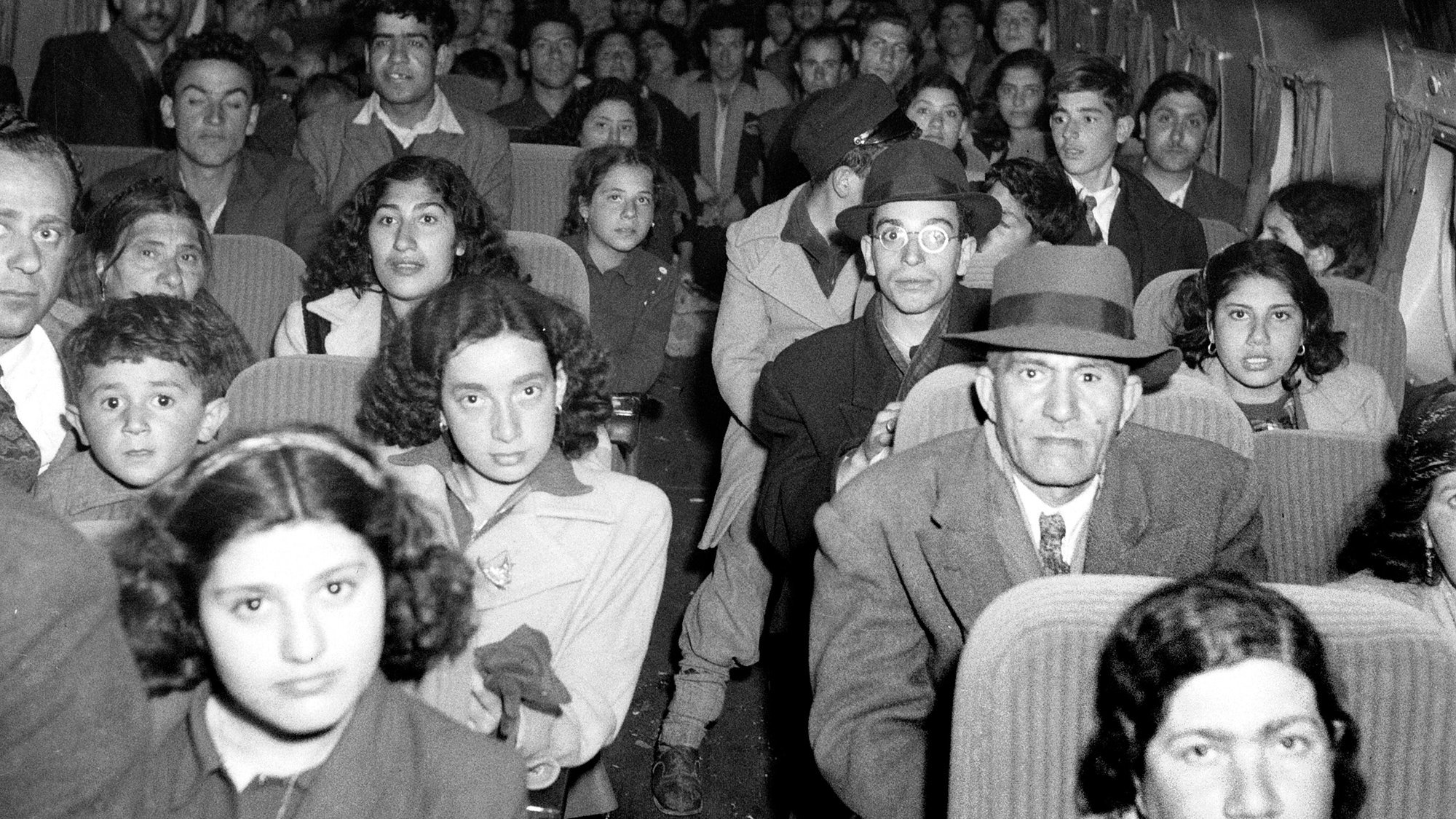 The image depicts a crowded interior of an airplane filled with passengers. There are men, women, and children seated in rows, looking directly at the camera. The individuals exhibit a mix of expressions, ranging from serious to curious. Many of them are dressed in clothing typical of earlier decades, suggesting that the photo may be historical. The cabin has a vintage appearance, and the layout and design of the seats reflect an earlier era of air travel.
