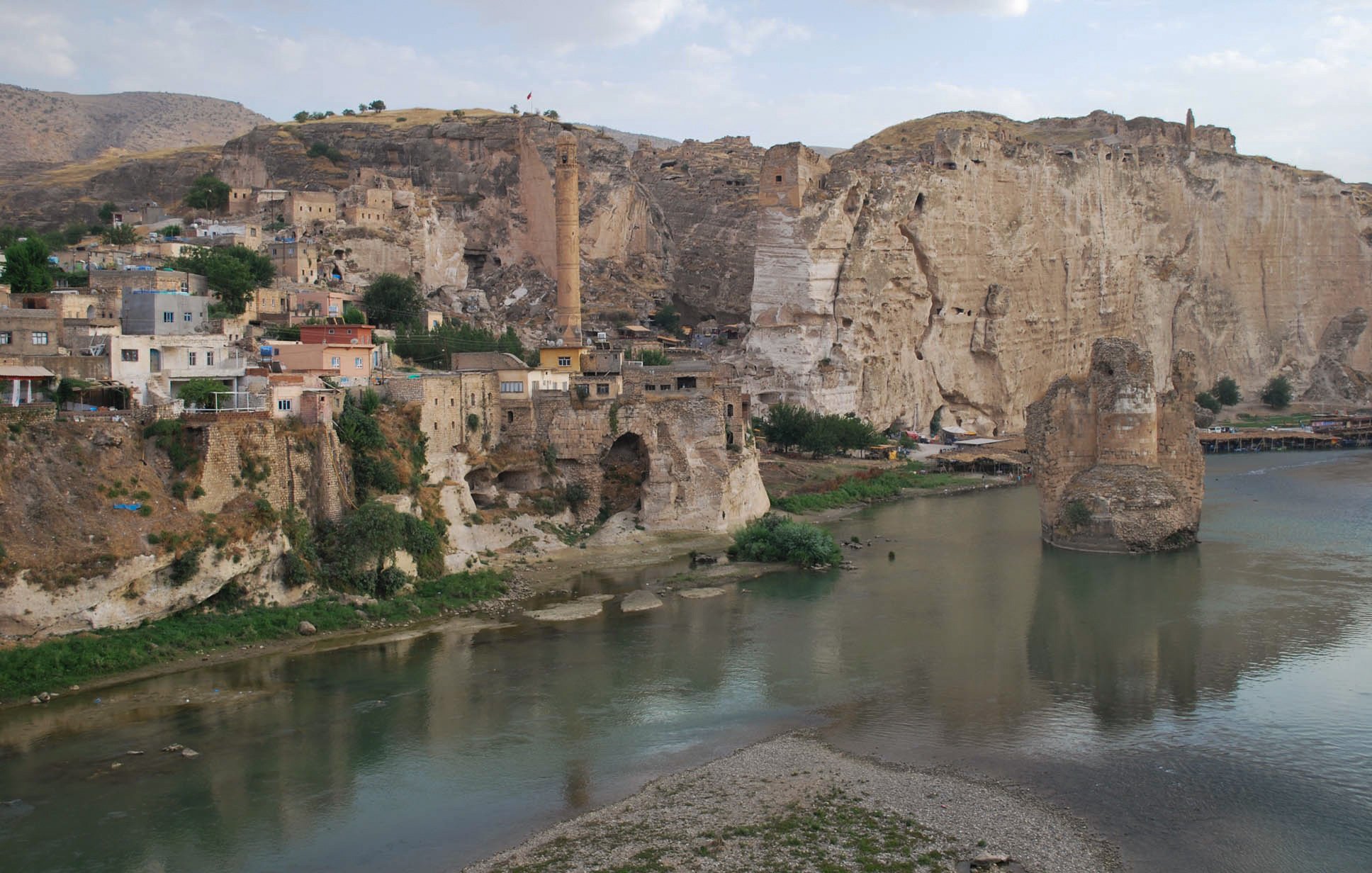 L'image montre un paysage pittoresque avec des formations rocheuses impressionnantes en arrière-plan. On y voit un village construit sur les flancs de la montagne, avec des maisons aux couleurs chaudes. Un fleuve calme coule au premier plan, reflétant les alentours. Quelques arbres verdoyants et des ruines anciennes ajoutent à la beauté historique de la scène. L'atmosphère est tranquille, dominée par les teintes naturelles des roches et de l'eau.