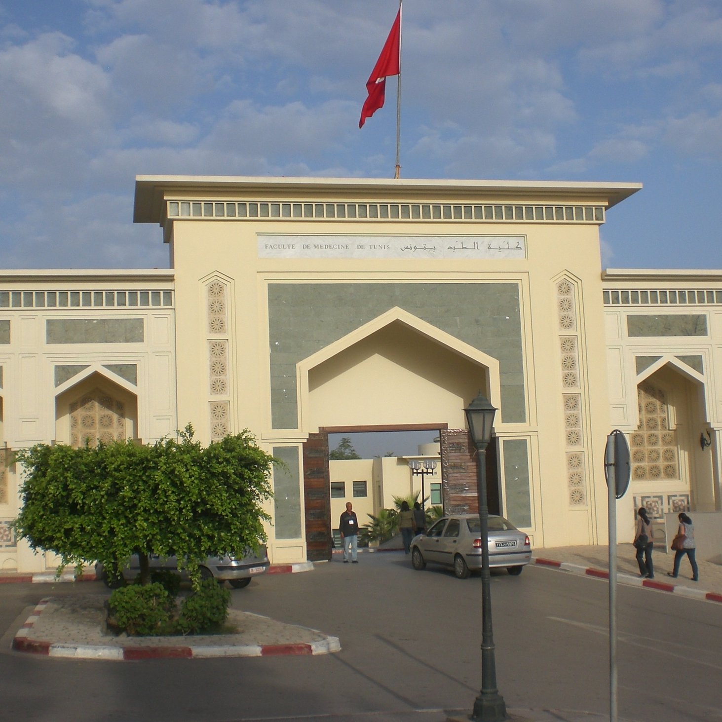 L'image montre l'entrée d'un bâtiment impressionnant, probablement une institution ou un musée, avec une architecture marocaine caractéristique. On peut voir une grande porte ornée de motifs traditionnels et des murs décorés avec des inscriptions en arabe et en français. Au-dessus de l'entrée, un drapeau rouge flotte. L'environnement est urbain, avec quelques arbres et des voitures stationnées. Le ciel est partiellement nuageux, apportant une atmosphère calme.