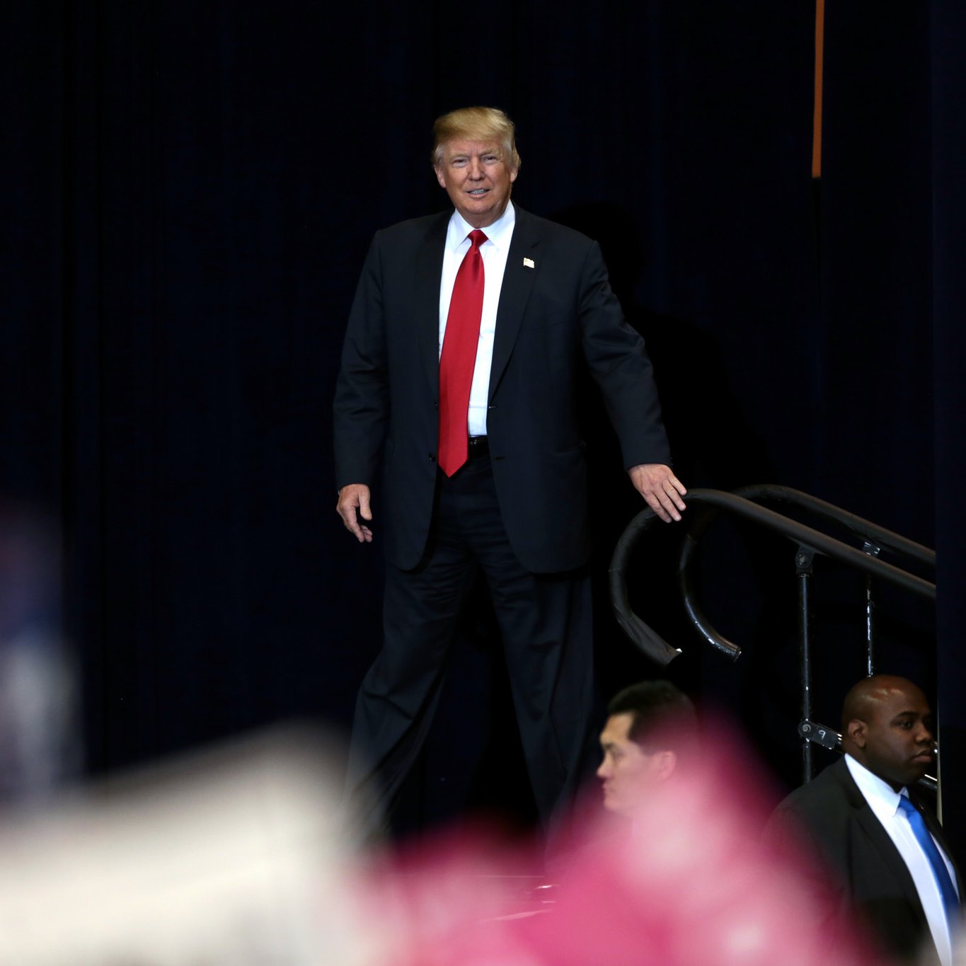 L'image montre une personne en costume, avec une cravate rouge, se tenant debout sur une scène. Il sourit et semble s'adresser à un public. Deux agents de sécurité se tiennent à proximité, surveillant la scène. L'arrière-plan est sombre, ce qui met en évidence la personne au premier plan. On voit également une foule en bas, bien que les détails soient flous.