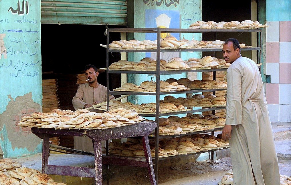 تُظهر الصورة مشهداً من أحد أسواق الخبز. يتواجد رجلان أمام رفوف مليئة بالأرغفة، حيث يبدو أن الرجلين مشغولان بعملية البيع أو التوزيع. الأرغفة مرتبة بشكل منظم على الرفوف، وتبدو طازجة. خلفهما، يمكن رؤية جدار ملون يُظهر بعض الكتابات. الجو يعكس أجواء الحياة اليومية في سوق محلي.