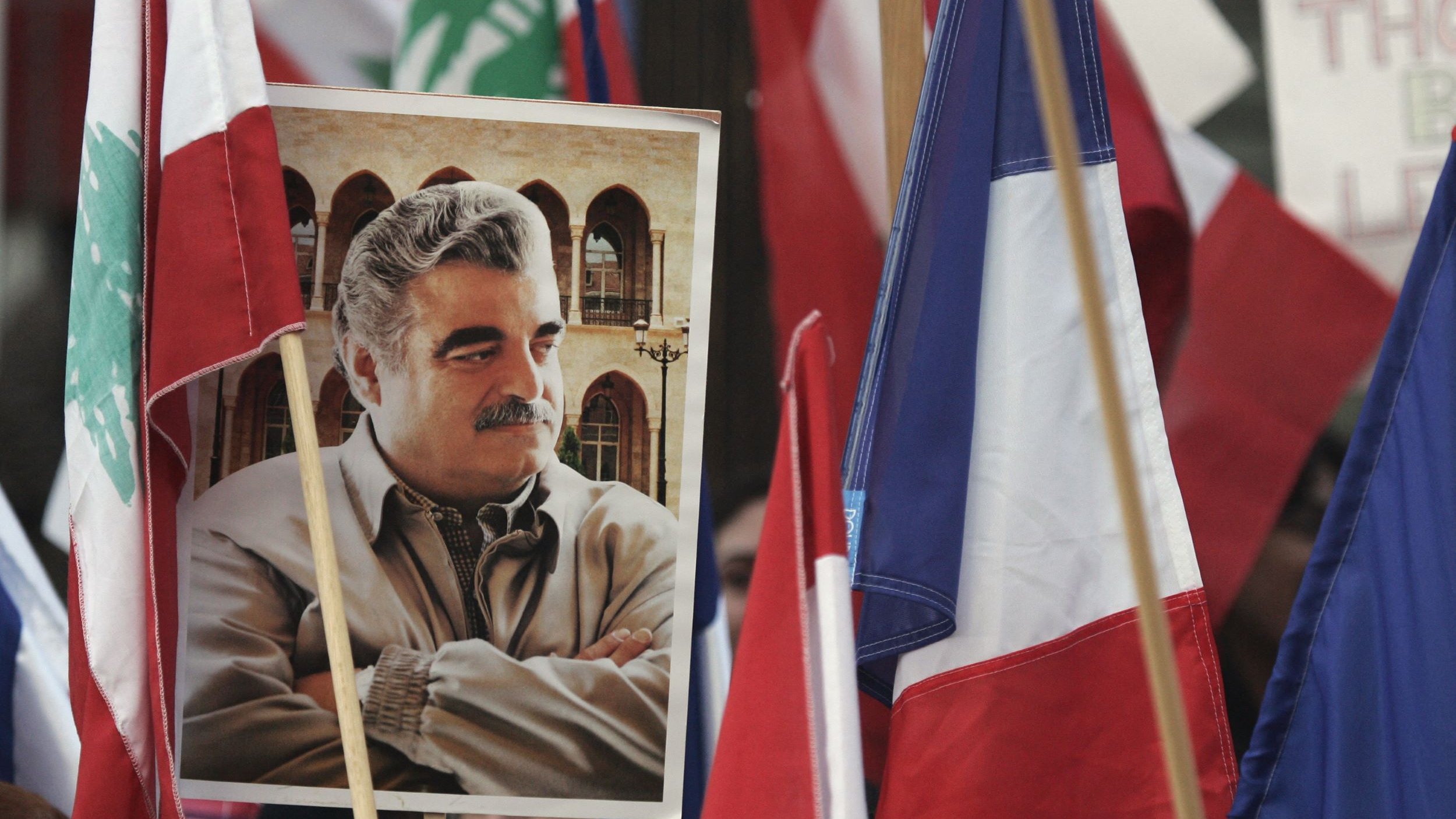 L'image montre une manifestation où plusieurs drapeaux, dont ceux du Liban et de la France, sont visibles. Au centre, il y a un portrait d'un homme, tenant une expression sérieuse, et entouré de personnes tenant des drapeaux. L'atmosphère semble chargée d'émotion et d'engagement.