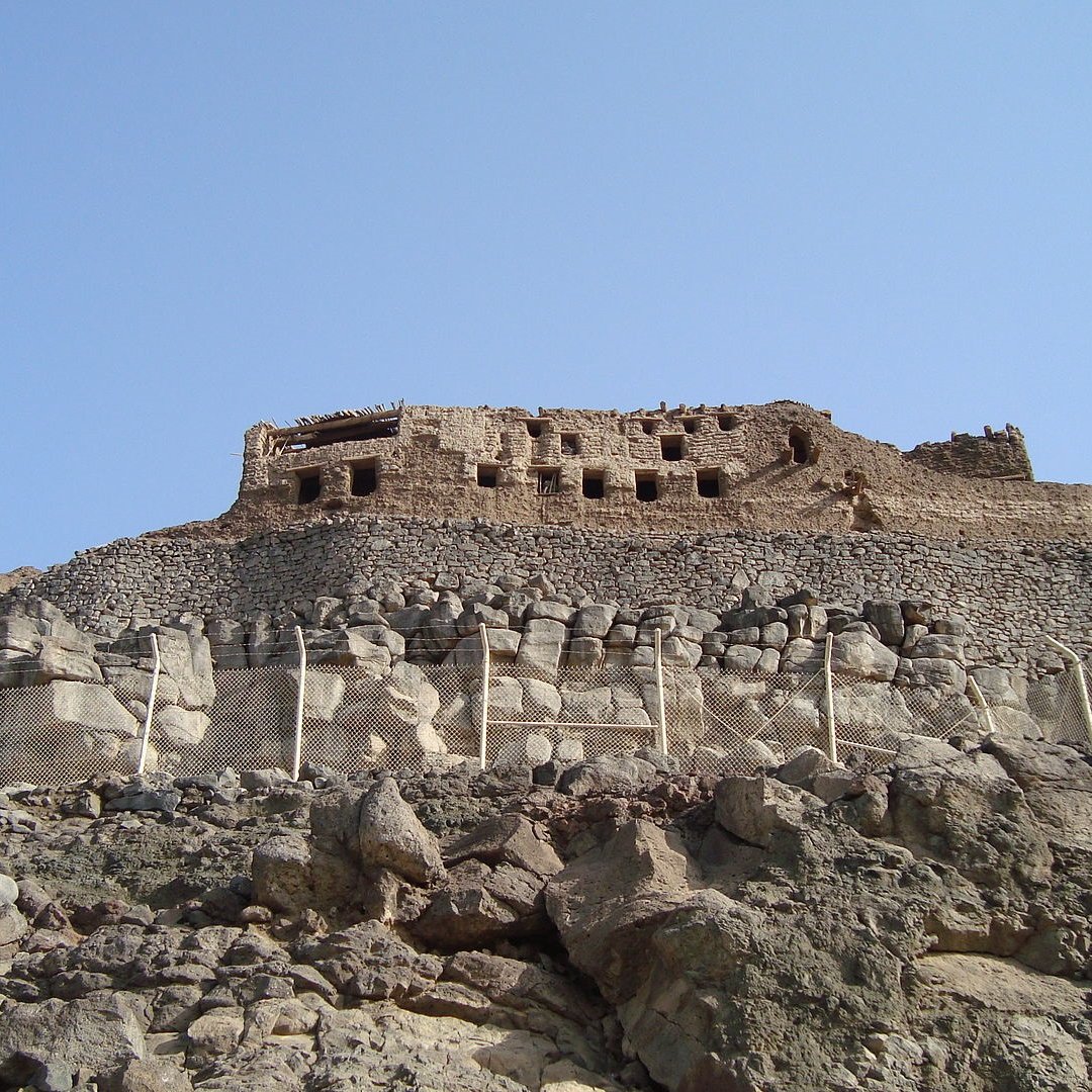 L'image montre une ancienne structure en pierre située sur une colline rocheuse. On peut voir des ruines dissimulées par des murs et des terrasses. Le ciel est dégagé, ce qui donne une belle luminosité à la scène. La végétation est absente, et le paysage est principalement composé de roches. La construction semble avoir une importance historique, suggérant qu'elle pourrait être un vestige d'une civilisation passée.