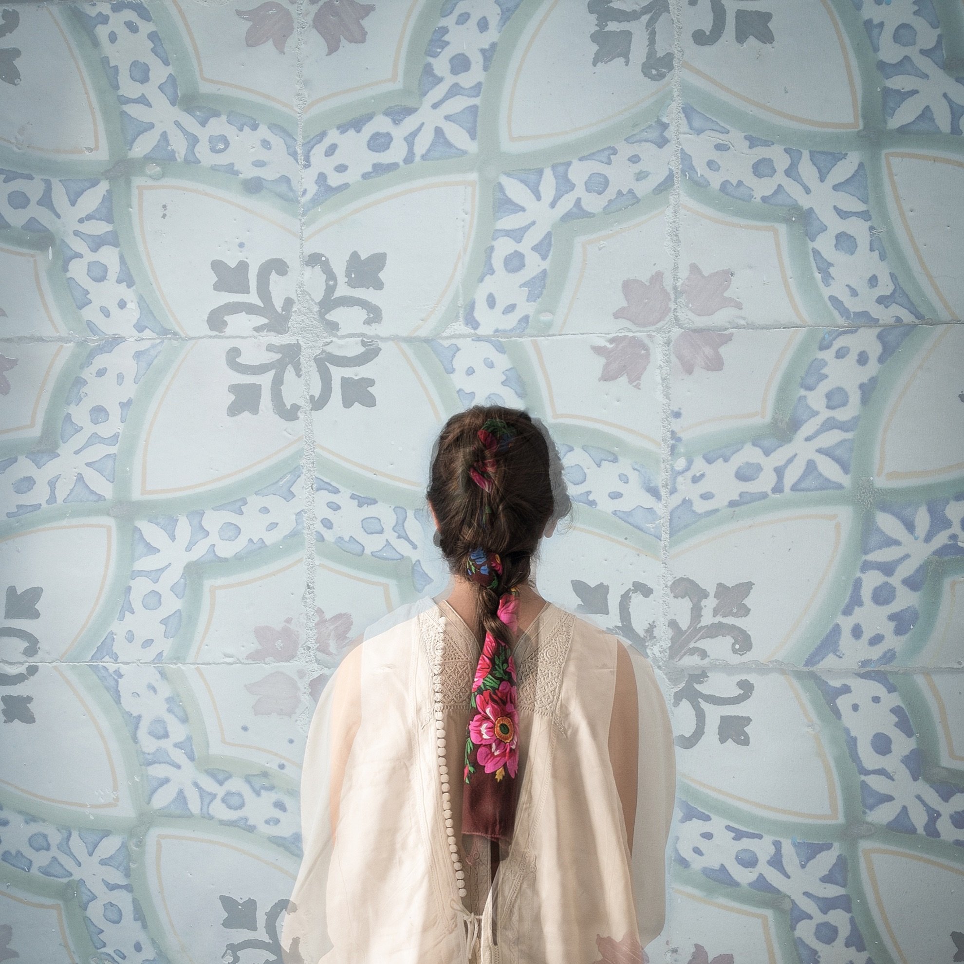 Cette image montre une personne de dos, portant un vêtement léger et délicat. Sa longue tresse, ornée de fleurs colorées, est visible et se détache sur un fond de motifs bleu clair. Les motifs sont ornés de formes géométriques et florales, créant une ambiance apaisante et artistique. L'ensemble évoque une atmosphère de calme et de beauté.